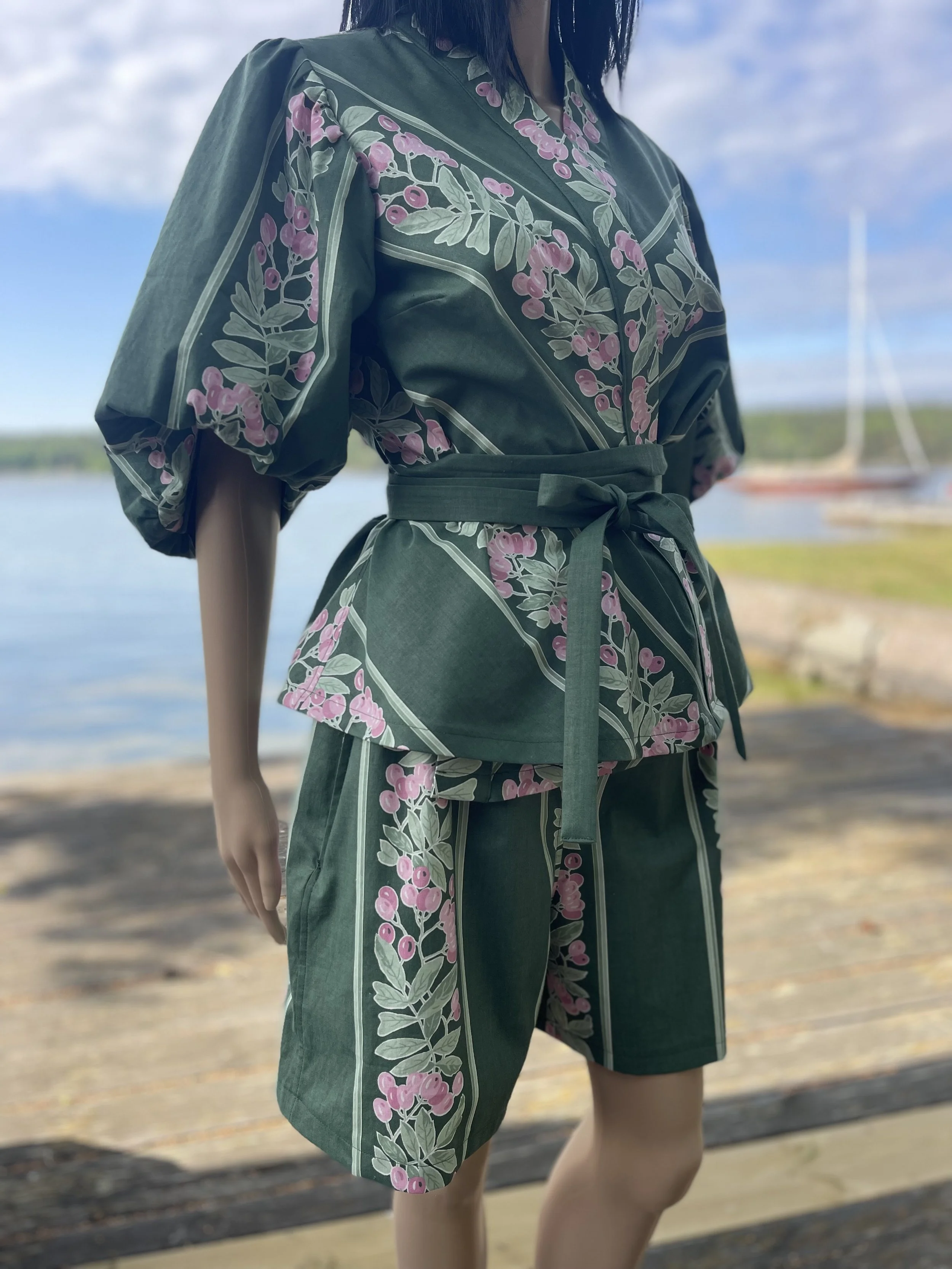 Woman wearing a matching green floral outfit with pink berries, standing near a body of water with an island and sailboat in the background.