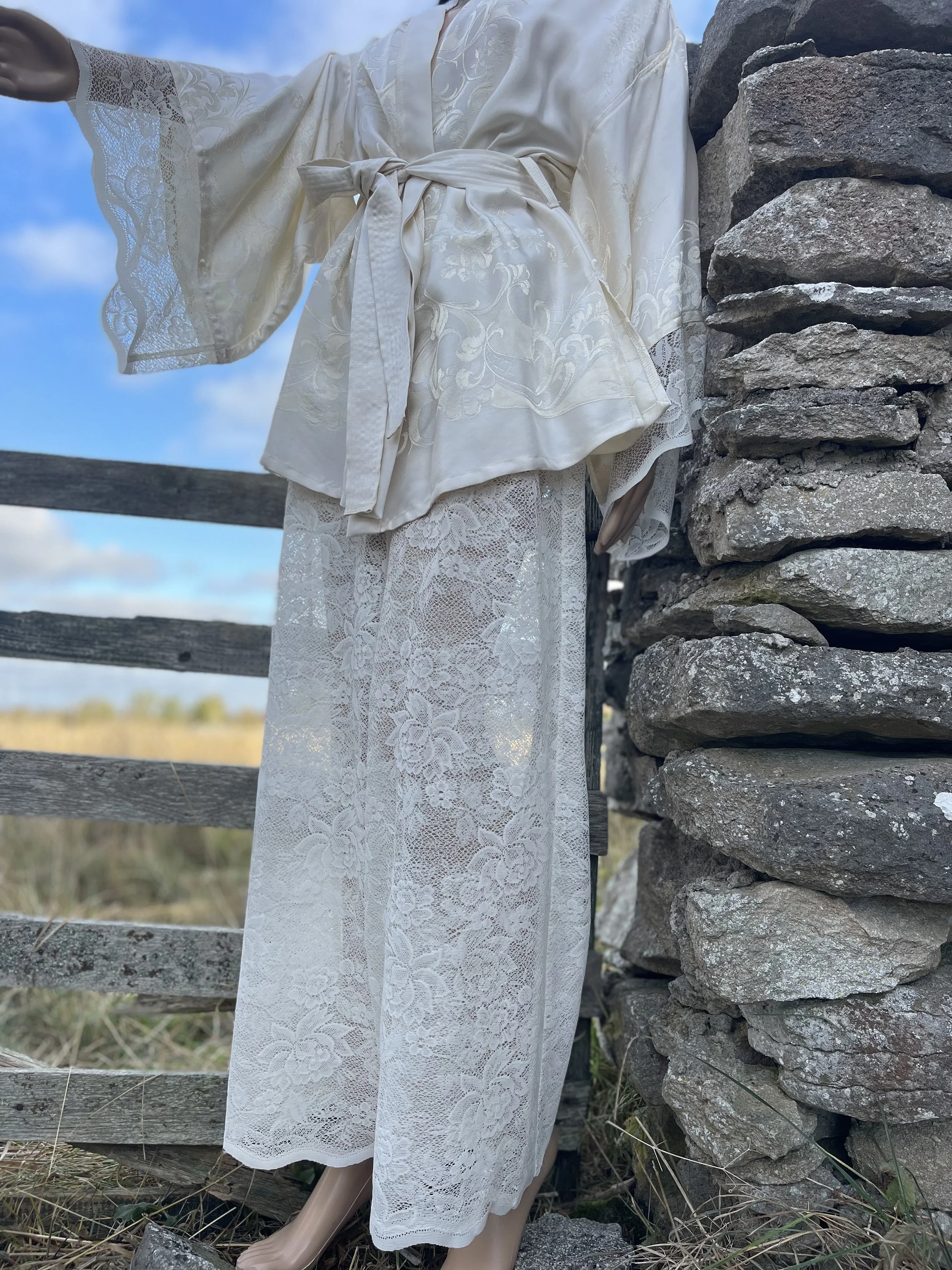 A mannequin dressed in a white lace and satin robe standing outdoors against a stone wall with a wooden fence and open sky in the background.
