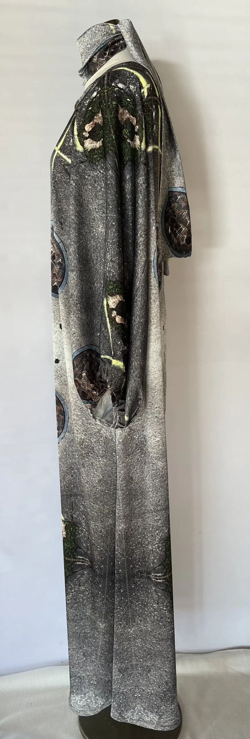 A mannequin dressed in a long coat and matching pants, both featuring a print of a gravelly ground surface with small patches of grass, and other patterned designs in dark tones.