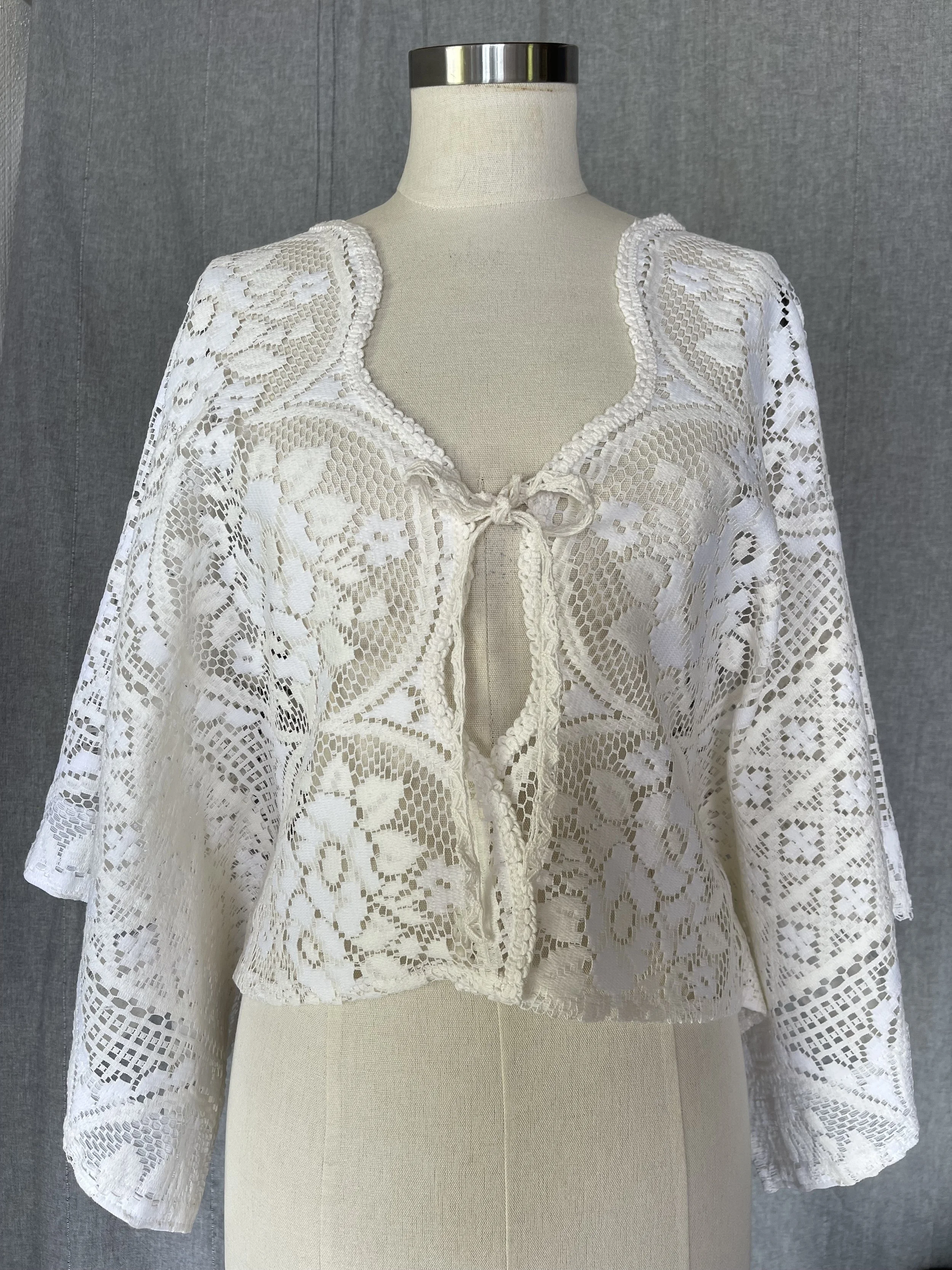 White lace blouse with floral pattern, long sleeves, and a front tie at the neckline displayed on a mannequin.