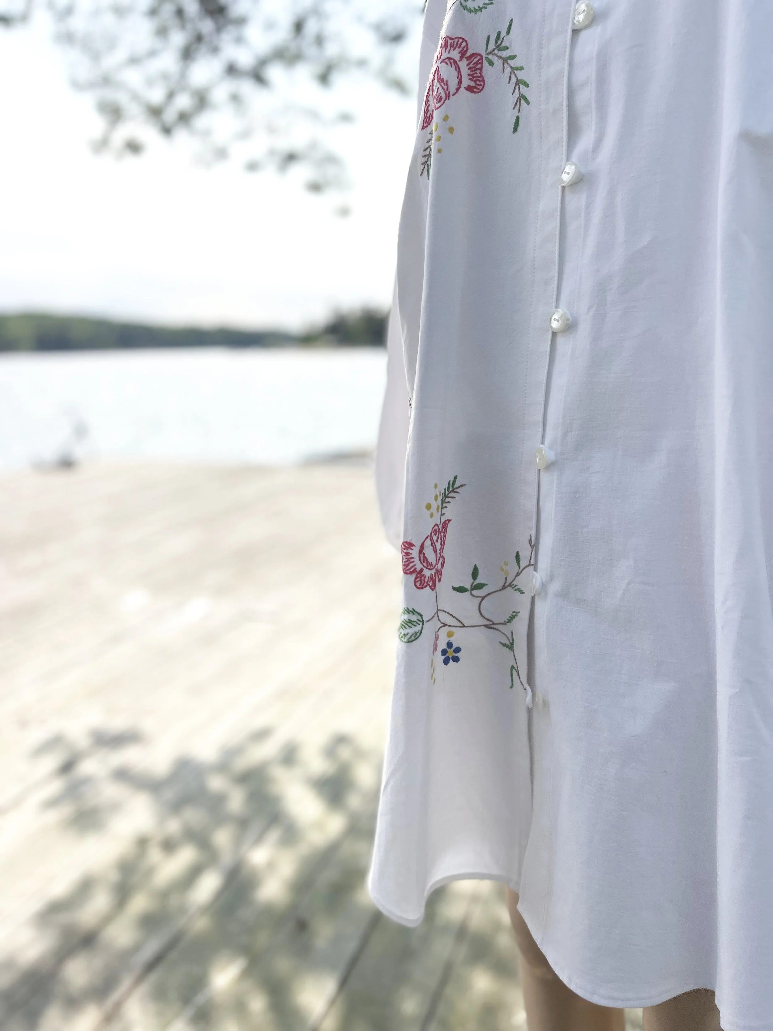 Close-up of a person wearing a white shirt with floral embroidery on the shoulder, standing outdoors near a body of water with trees in the background.