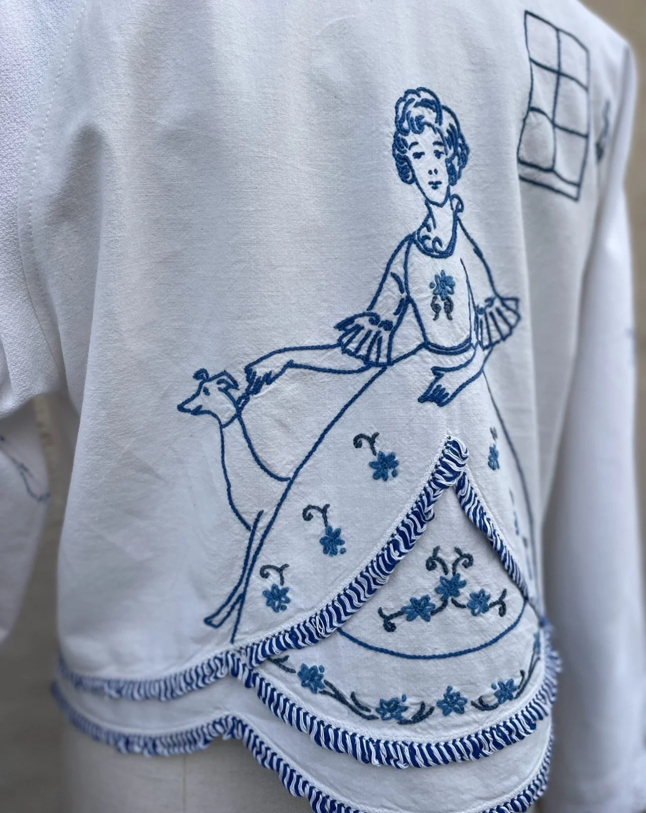White fabric with blue embroidery of a woman in a dress, holding a small dog, with a window in the background.