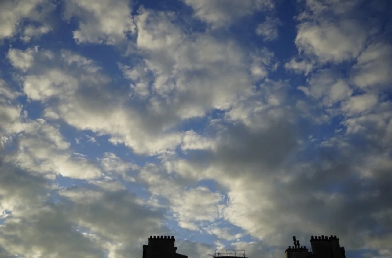 Sky filled with scattered clouds above dark silhouettes of rooftops with chimneys.