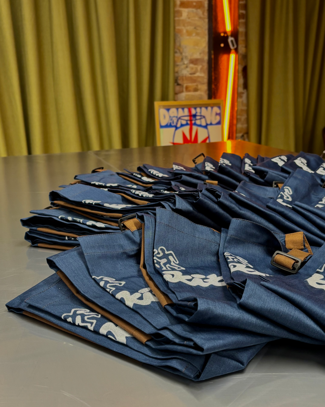 Multiple dark blue drawstring bags with white logos laid out on a table in a room with green curtains and a neon sign in the background.