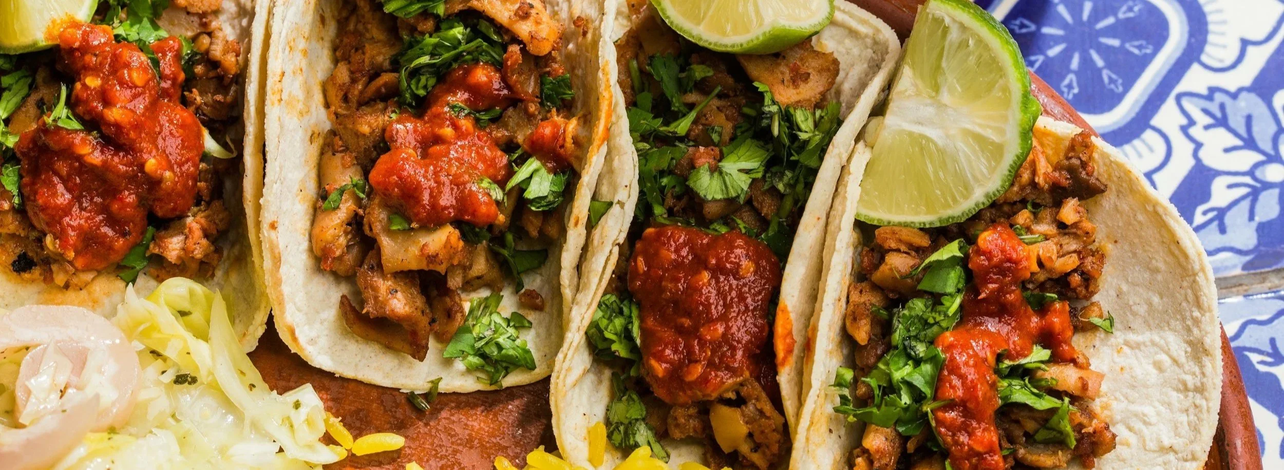 Zbliżenie na trzy tacos z wołowiną, kolendrą, kawałkami limonki i salsą na talerzu w niebiesko-białe wzory.