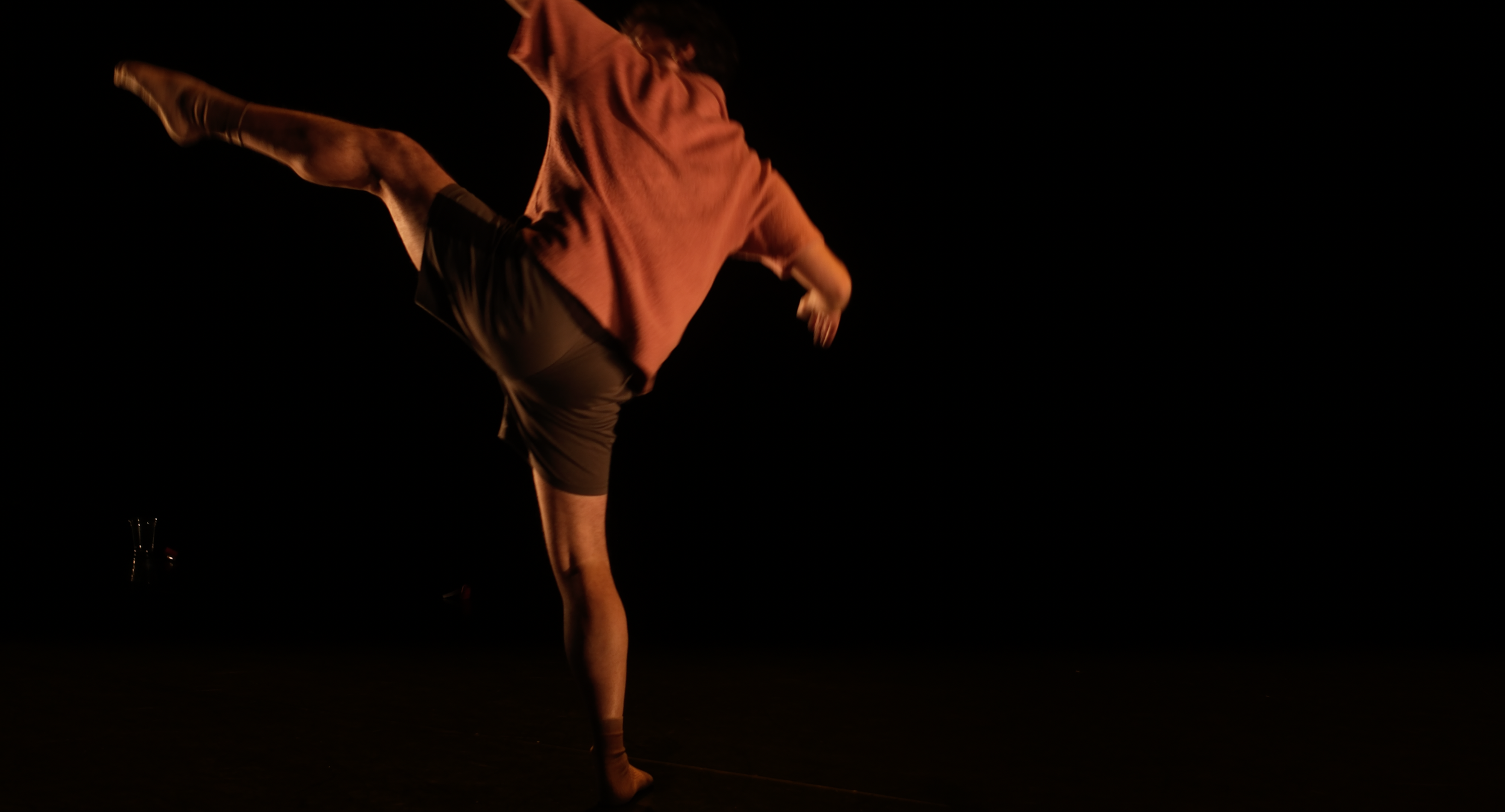 Aaron is seen facing in pitch black, facing the back of the stage with one arm and leg raised high into the air. 