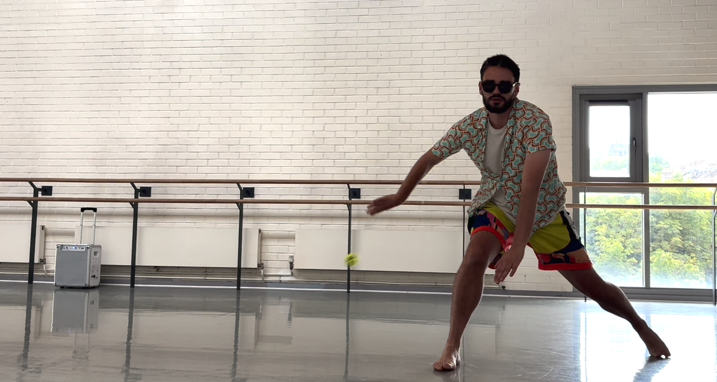 Aaron is seen in a wide pose, wearing sunglasses and colourful outfit, flinging a tennis ball across the room.