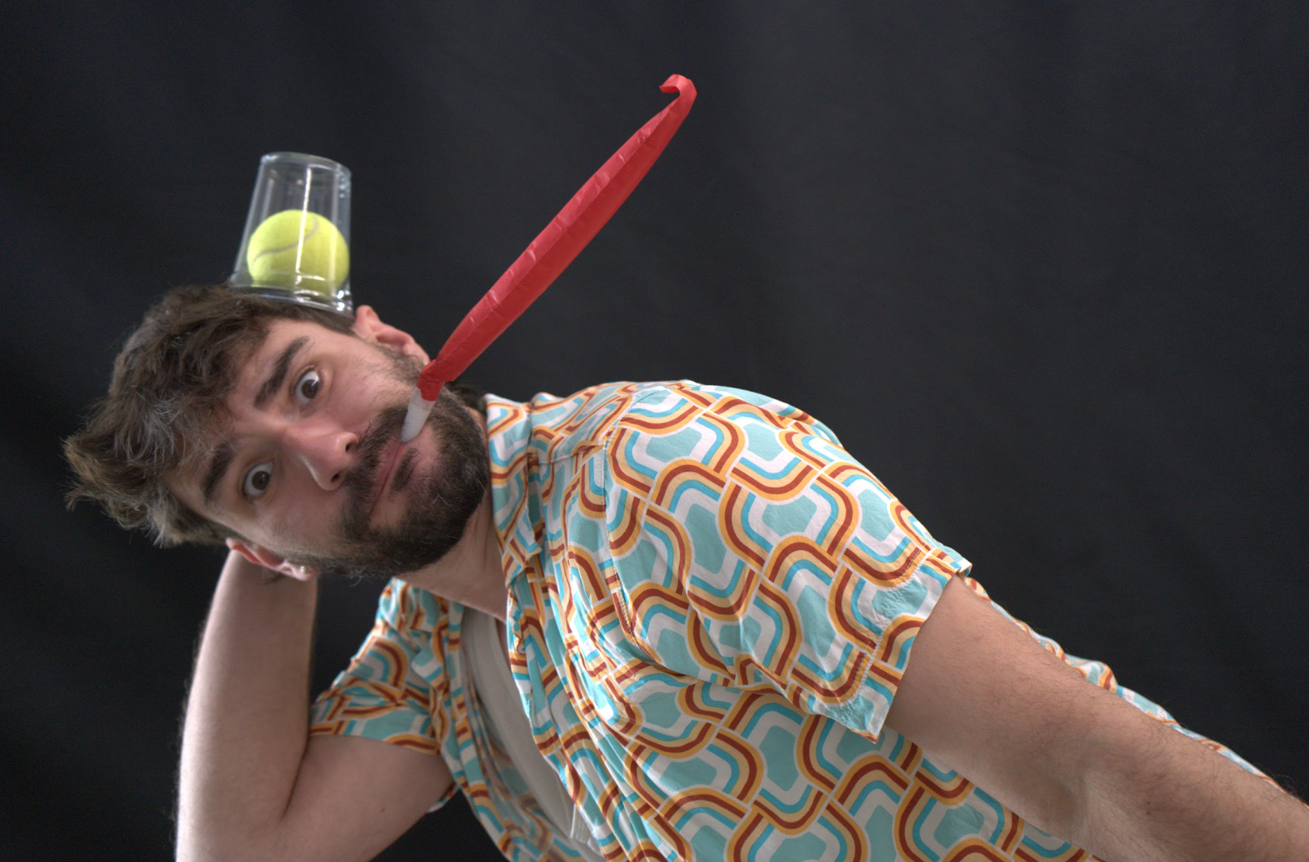 A male performer (Aaron Markwell) is seen tilting to one side, blowing a red party streamer with a tennis ball and cup balanced on his head.