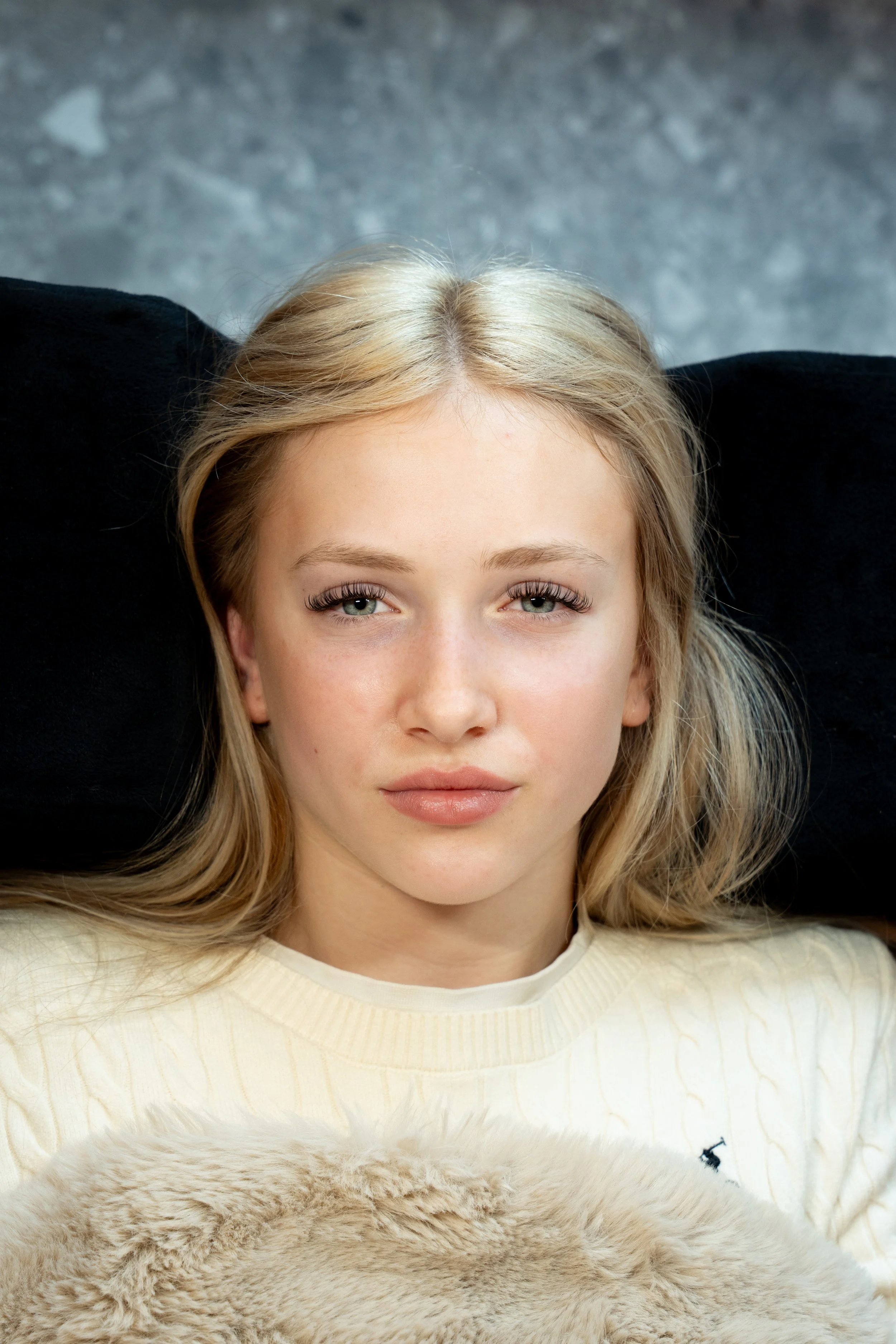 Close-up van een jonge vrouw met blonde haar die op haar rug ligt, haar hoofd rust op een zwarte kussen, met een grijze vloer op de achtergrond.