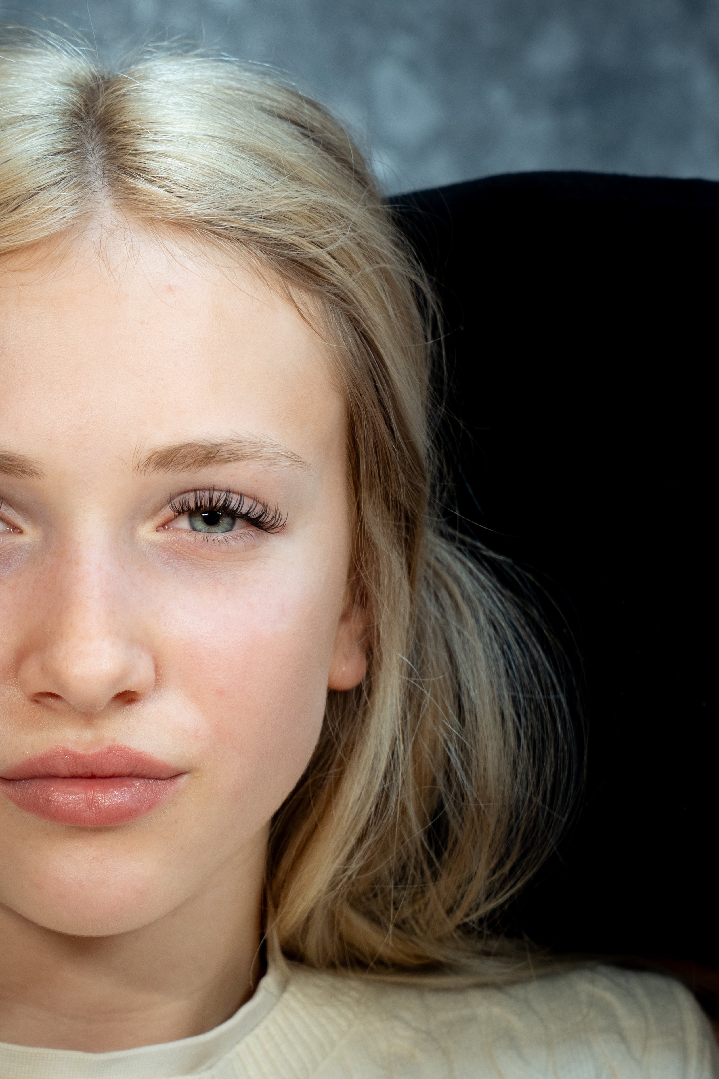Close-up van een jonge vrouw met lichte huid, blond haar, blauwe ogen en een lichte glimlach, tegen een donkere achtergrond.