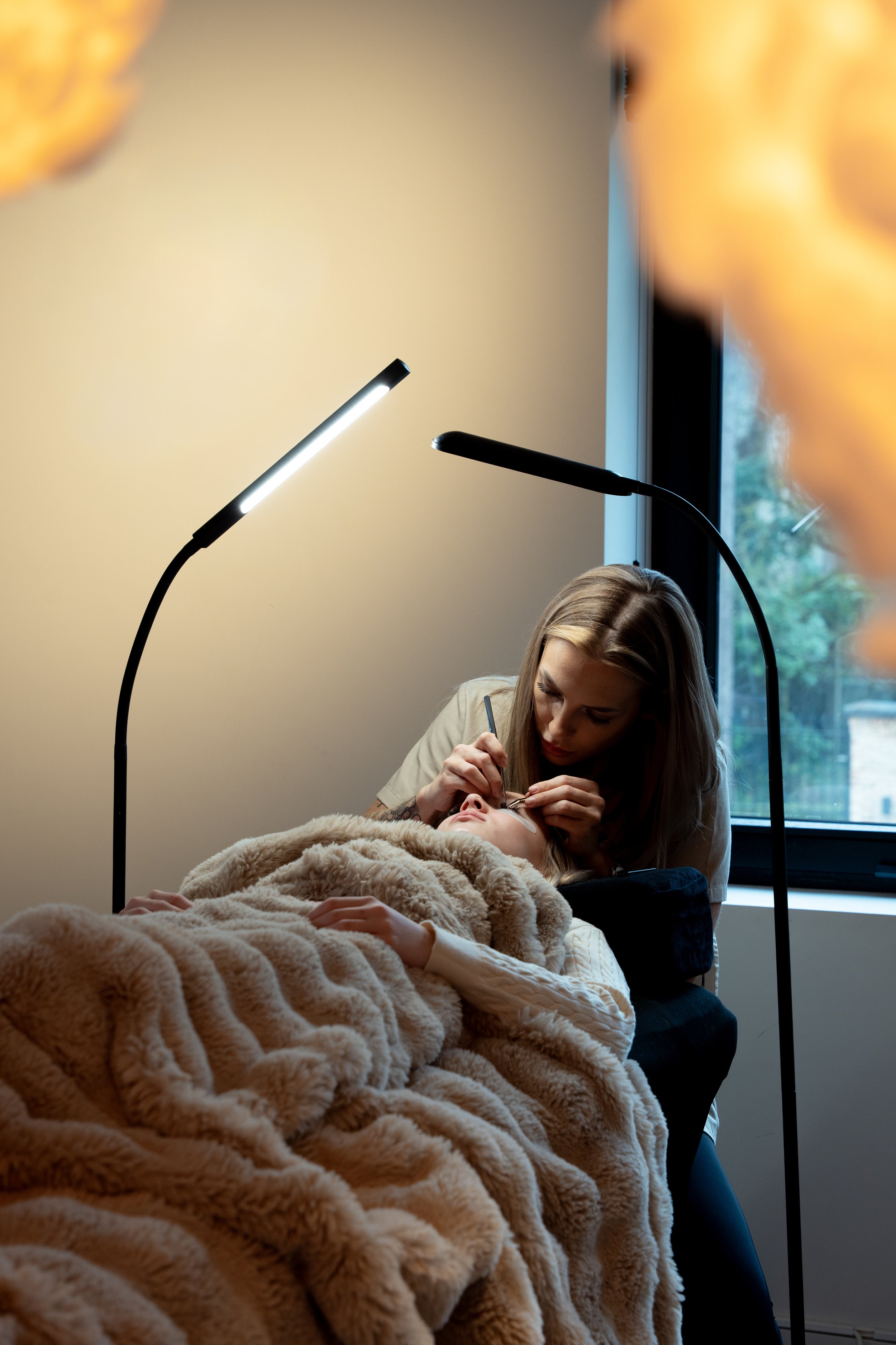 Een vrouw krijgt make-up aangebracht op een jonge dame die op een kliniekbed ligt, met een beige deken erover.