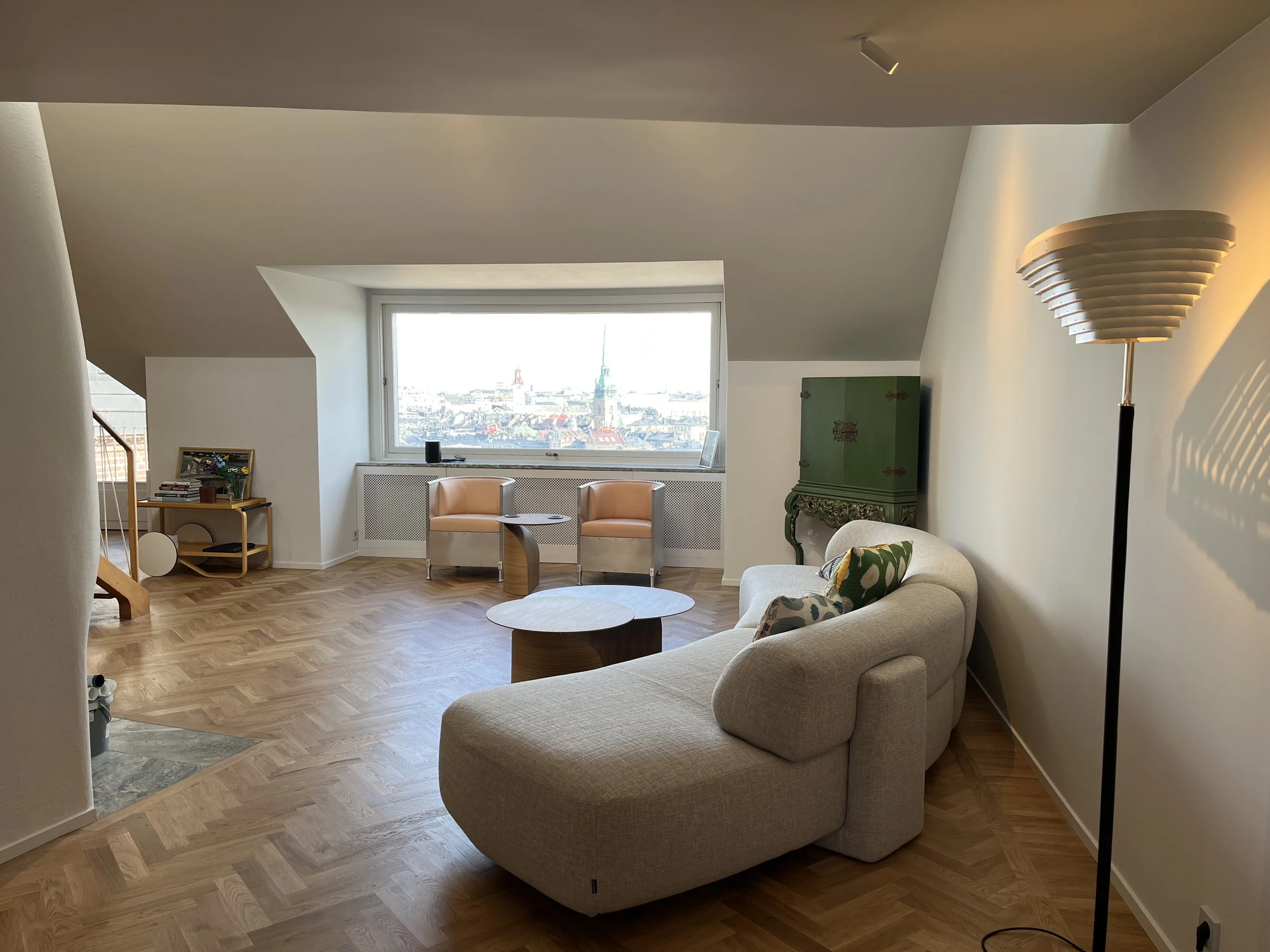 Ett vardagsrum med sittplats, ett stort fönster med utsikt över staden, ett grönt skåp, en luftpumpad golvlampa och ett soffbord.