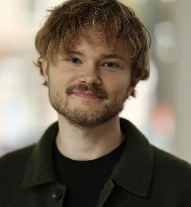 En ung arkitekt med lockigt, ljust hår och lätt skägg, bär en mörk jacka och ler mot kameran.