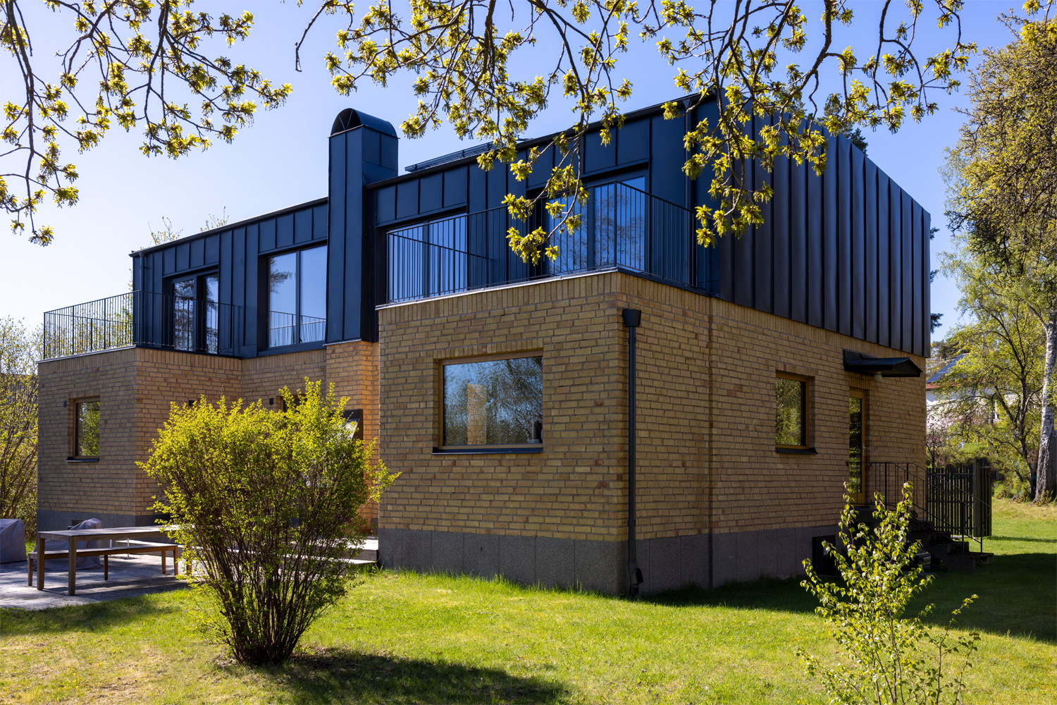 Modern villa med två våningar, gul tegelstenar och svart plåttak, omgiven av gröna träd och gräsmatta, under en klarblå himmel.