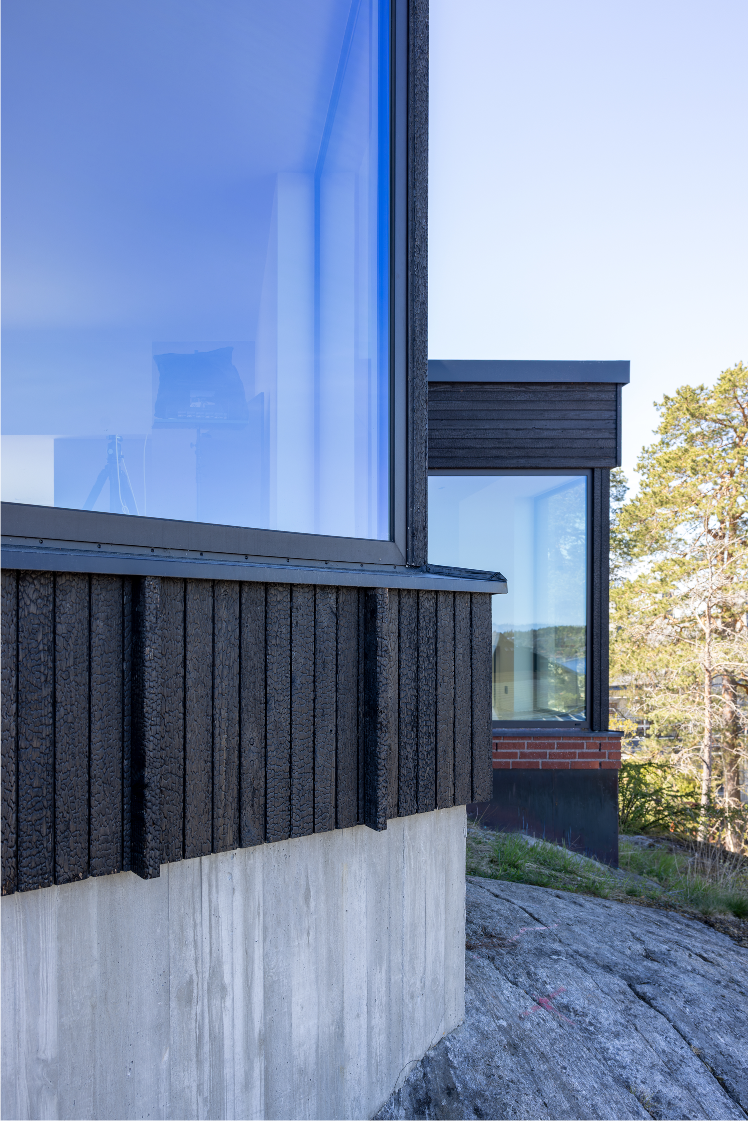 Närbild av ett modernt byggnadskomplex med stora glasfönster och svart träfaçad, beläget i ett grönt område med träd i bakgrunden, under en klar blå himmel.