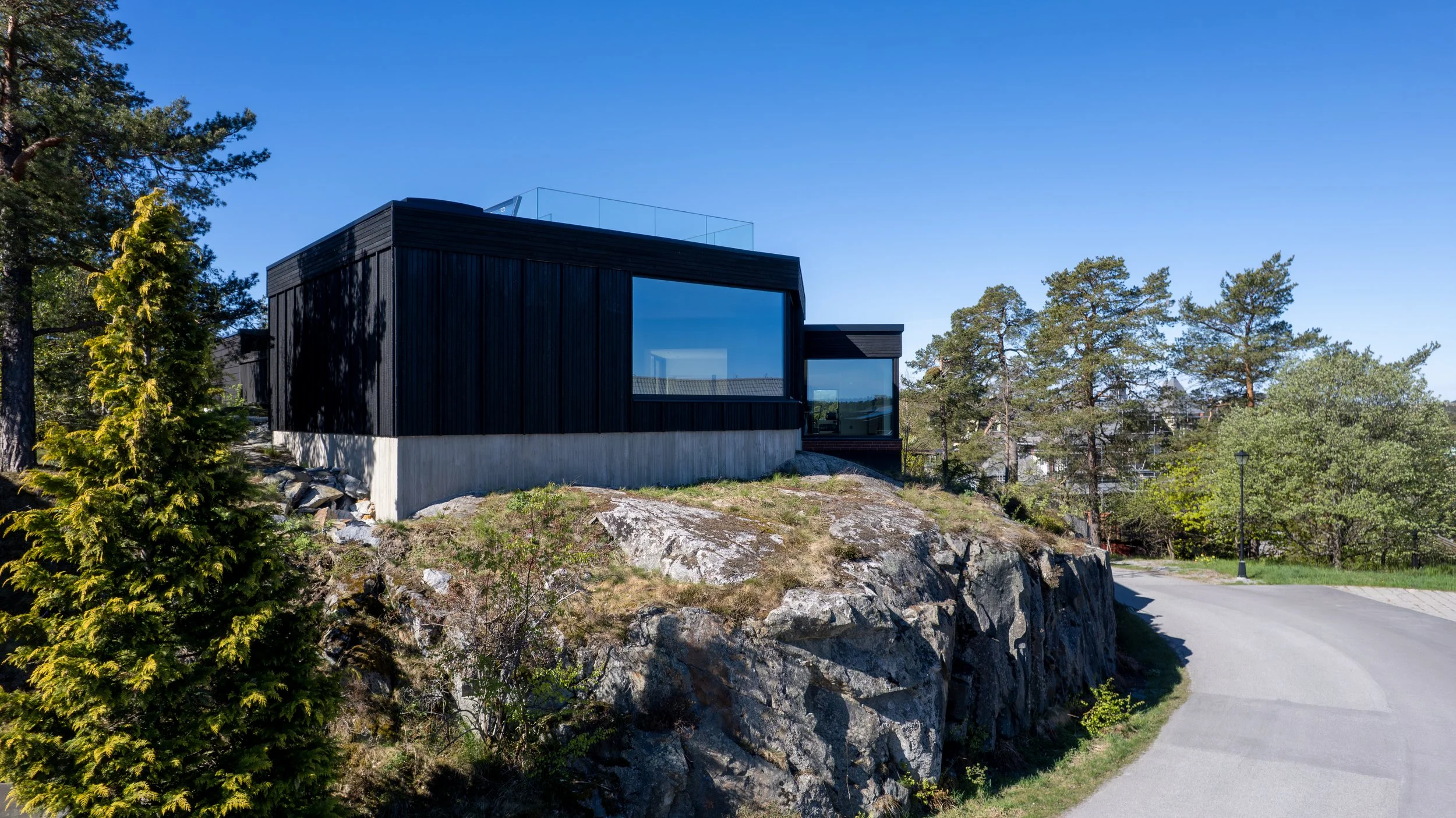 Modern svart klädd villa med stora fönster på en bergig plats, omgiven av träd och en grusväg under en klar blå himmel.