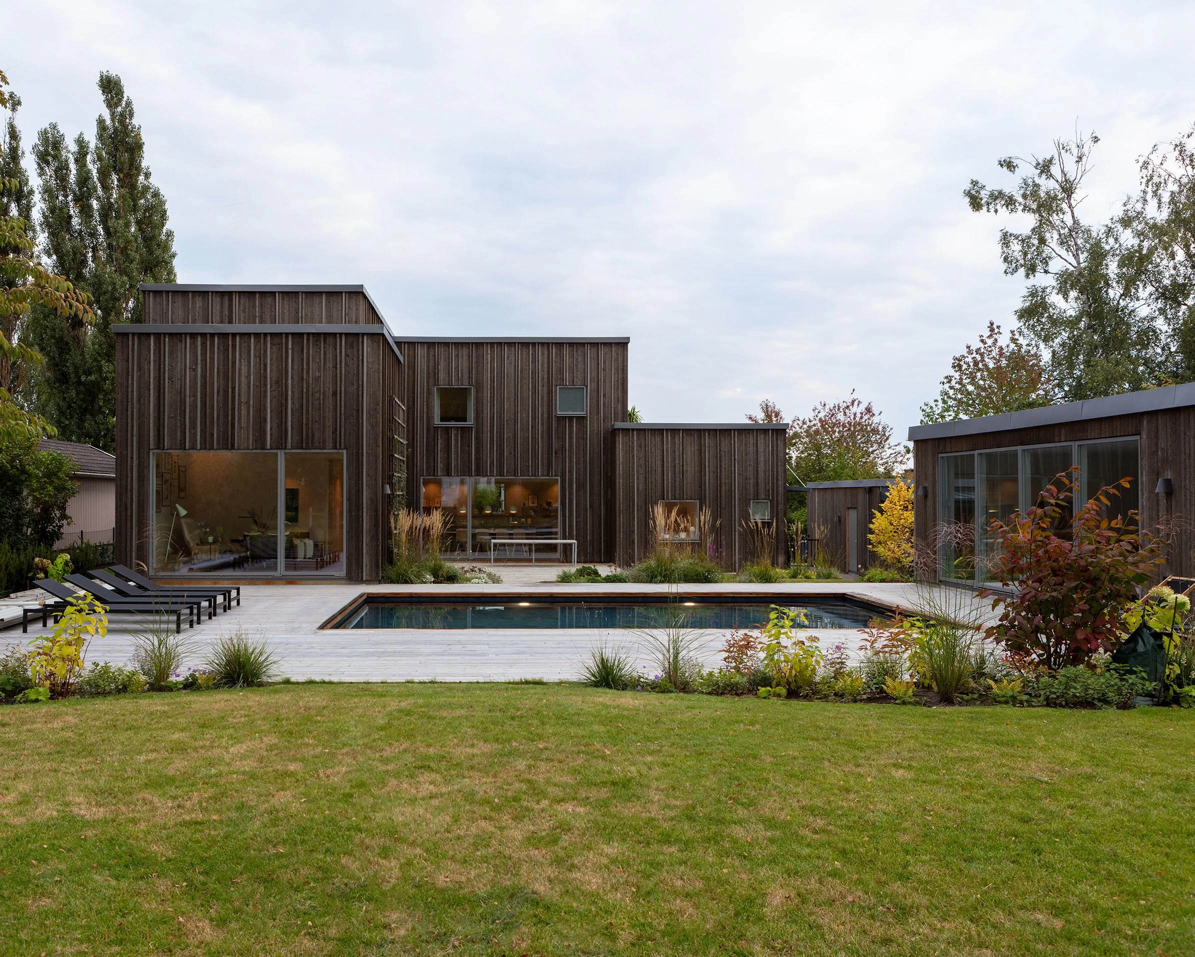 Modern träbyggnad med stoat fönster mot en trädgård och pool, omgiven av träd och buskar, under en molnig himmel. Ny villa i Näsby Park, Täby.
