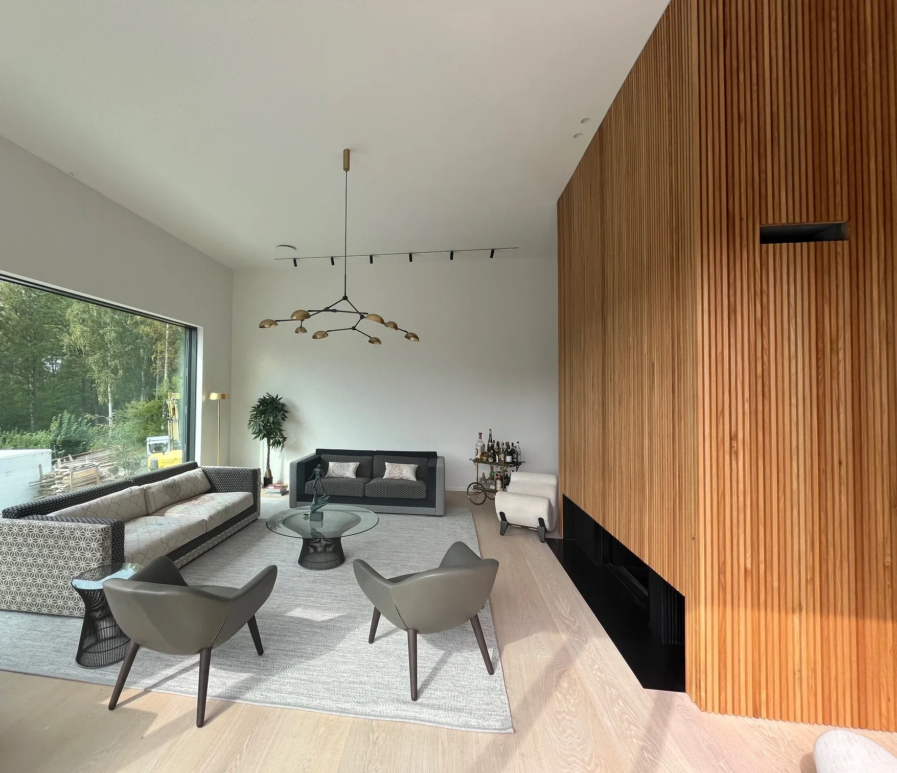 Vardagsrum med stora fönster mot trädgård, soffor, fåtöljer, vägghängd lampa, trävägg och moderne lampor.