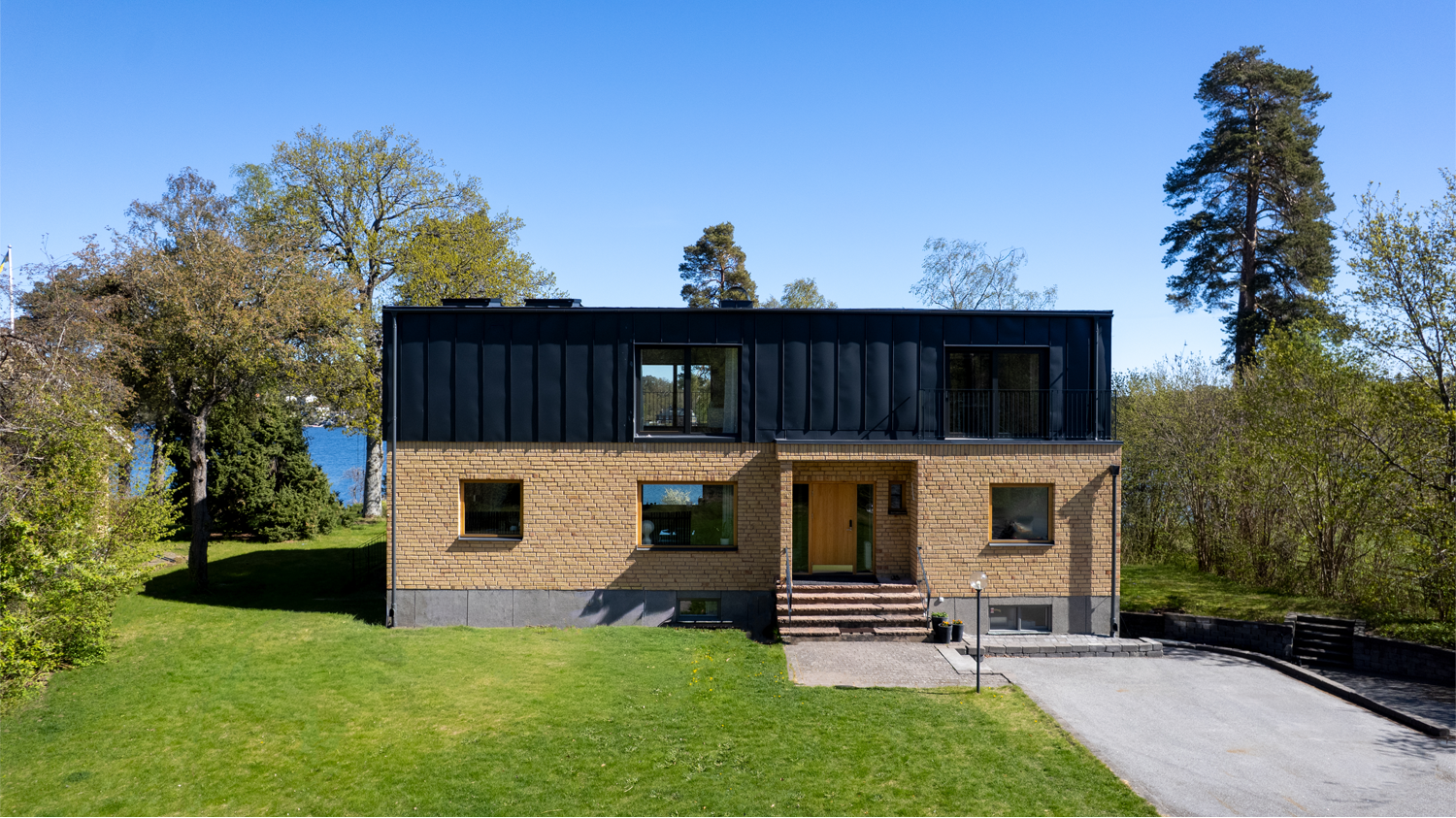 Modernt hus med tegel på nedre plan och svart metallisk övervåning, framför en grön trädgård med trappsteg och en grusstig, omgiven av träd och blå himmel.