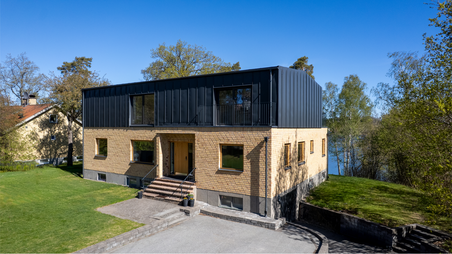 Villa med två våningar, gult tegel på nedre och svart metall på övre våningen, omgiven av grönt gräs och träd, med en liten trätrappa vid entrén