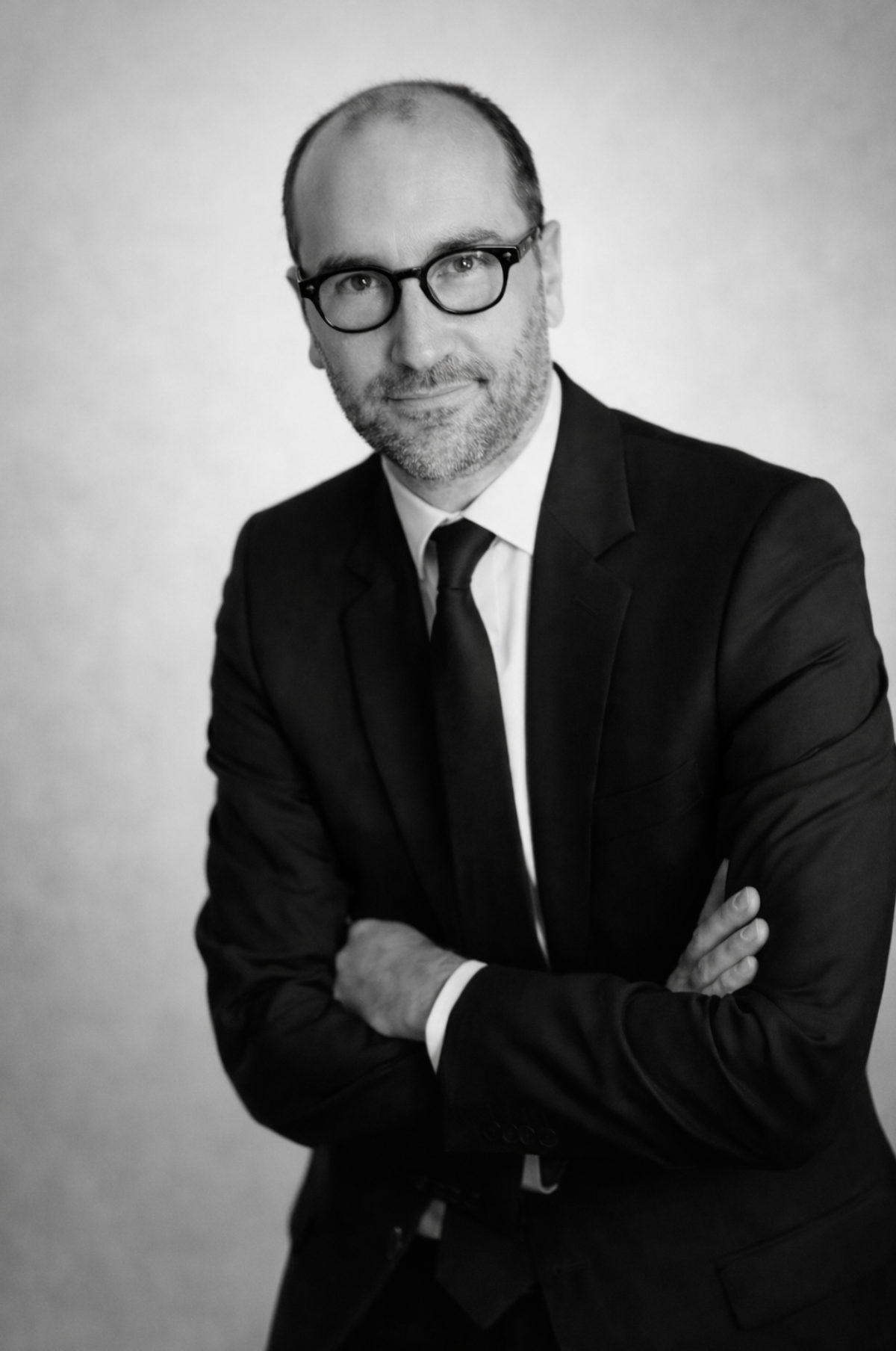 Portrait en noir et blanc d'un homme d'âge moyen portant des lunettes, en costume noir avec une chemise blanche et une cravate noire, bras croisés, regardant l'objectif avec un léger sourire.