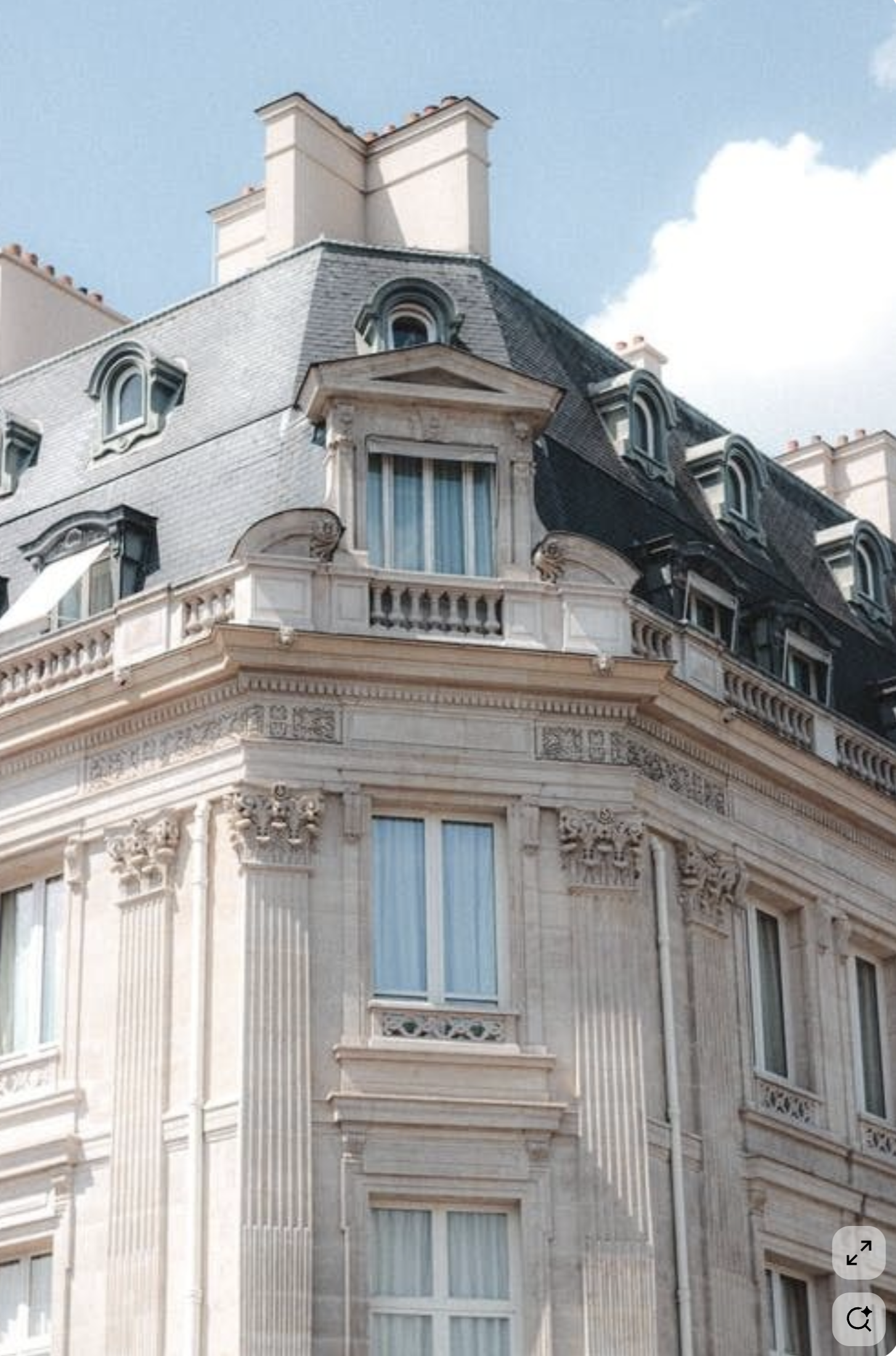 Un immeuble de style haussmannien avec des fenêtres à volets bleus, des balcons en pierre et une toiture en ardoise, sous un ciel bleu avec quelques nuages.