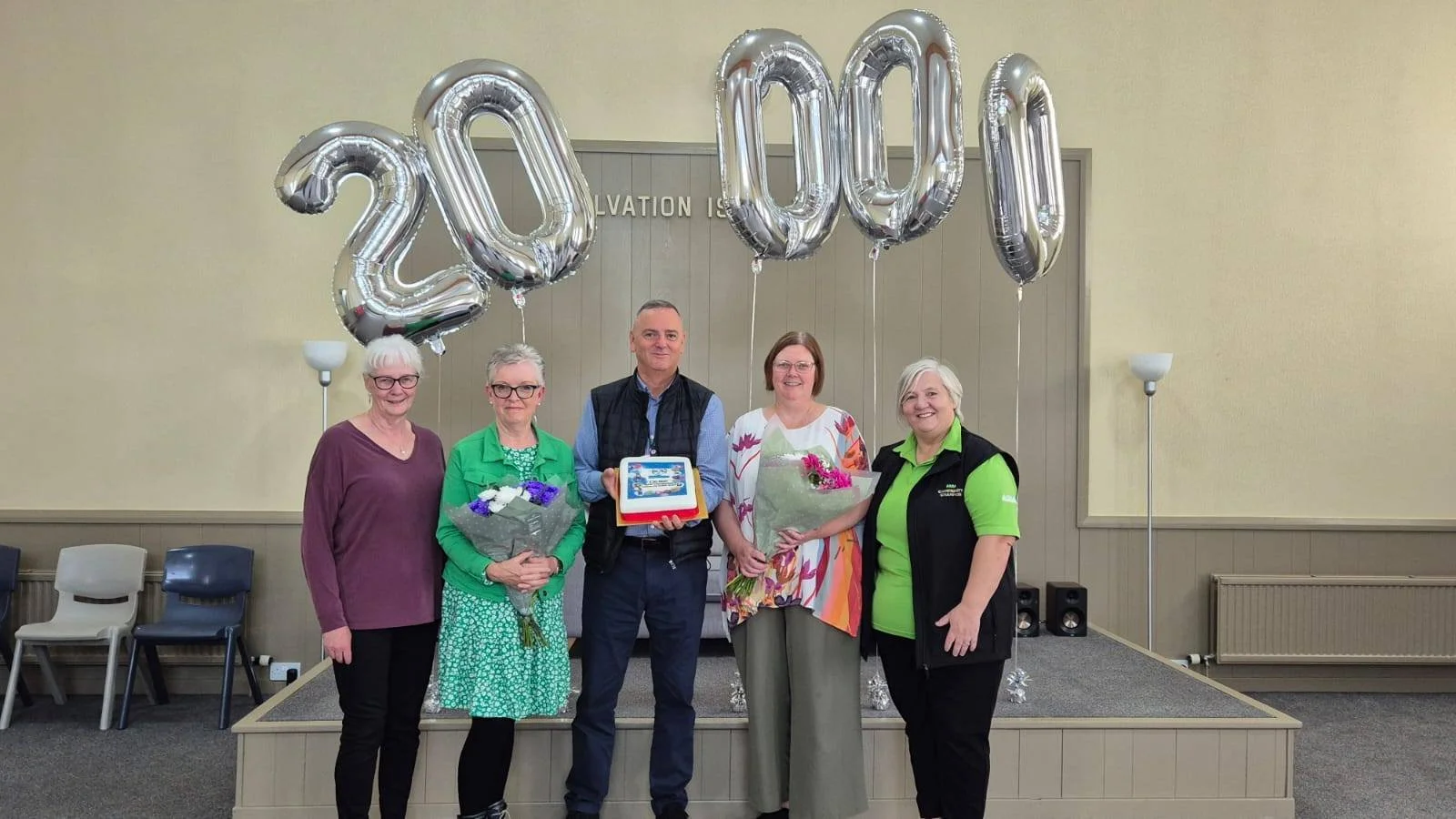 Five people standing on a stage with large silver balloons forming the numbers 2020 behind them. The group includes four women and one man, some holding bouquets of flowers. The man in the center is holding a cake. They are smiling, and the setting appears to be a celebration or recognition event.