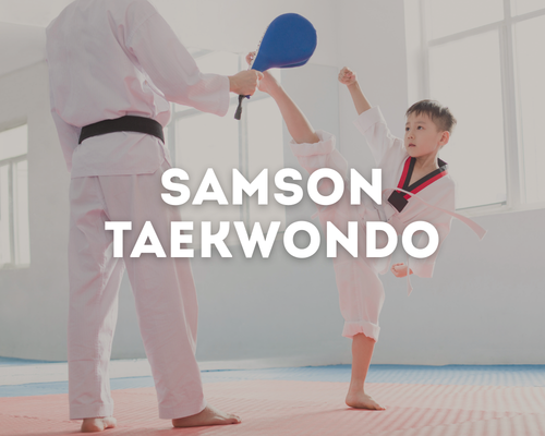 Young boy in martial arts uniform practicing taekwondo with instructor holding a blue paddle in a bright training room.