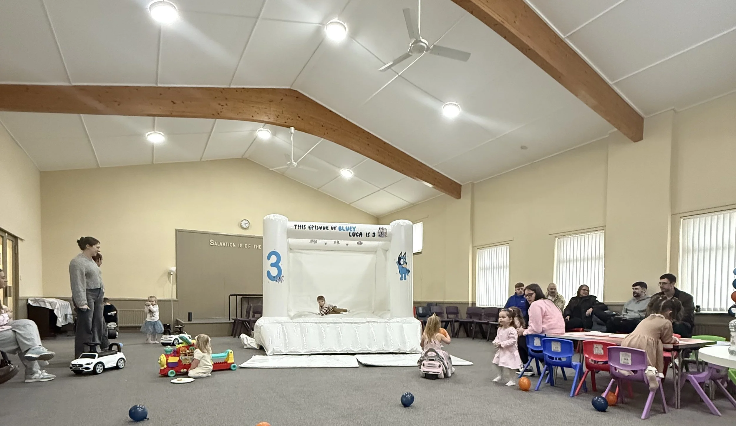 Children and adults in a large indoor room with beige walls, grey carpet, and a high ceiling with exposed wooden beams. Kids are playing on an inflatable slide at the center, while older individuals sit on chairs along the wall, and some children are