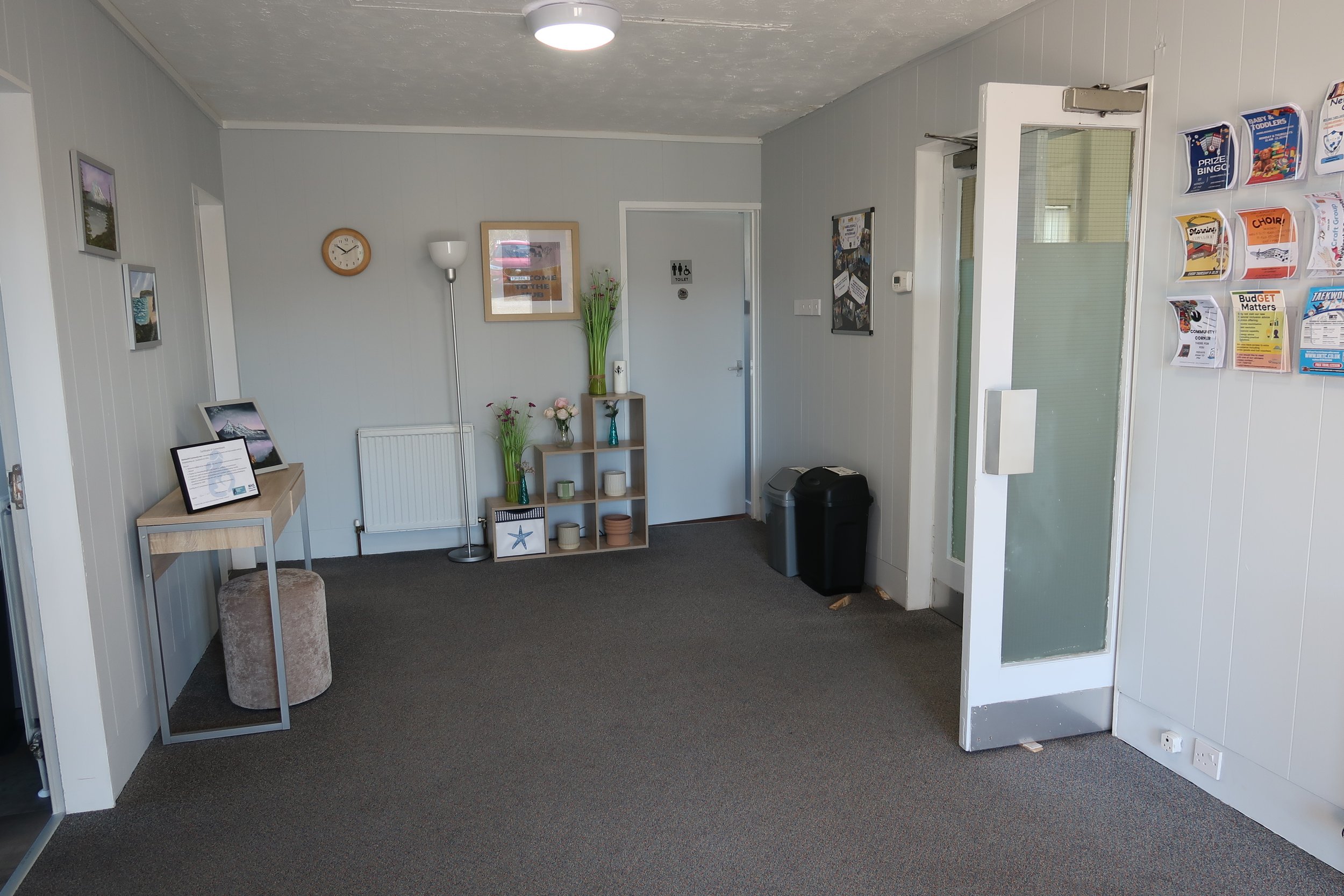 Interior view of a waiting room or reception area with a gray carpet, white paneled walls, and decorative items including framed pictures, a clock, vases, and brochures. There is a small table with a framed document, a stool, a tall floor lamp, a shelving unit with flowers and decorative objects, heating radiator, and trash bins. A door labeled 'Toilets' is visible, with a partition and brochure rack on the wall.