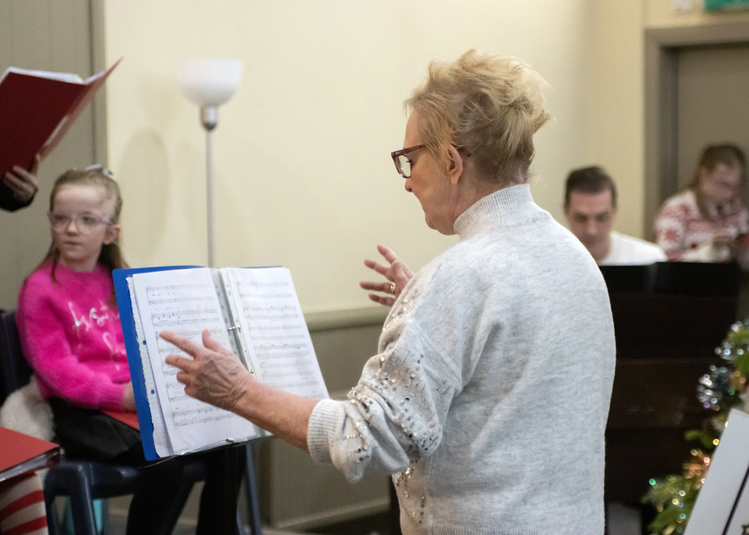 An elderly woman conducting a choir of children and adults in a music rehearsal or choir practice, with music sheets in front of her and other participants in the background.
