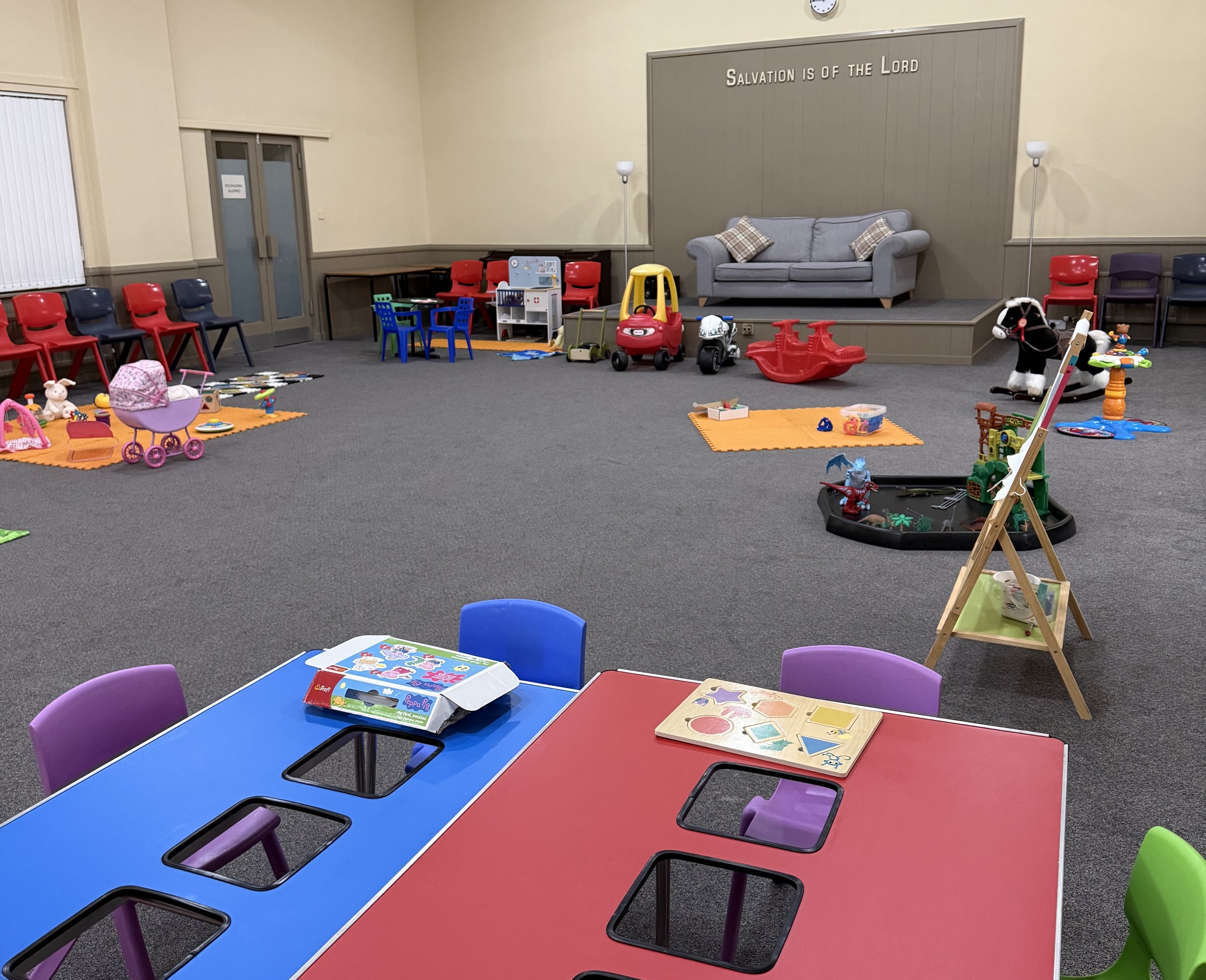 Empty children's playroom and activity area with toys, chairs, and a stage with a sofa, displaying the setup for Baby & Toddlers group.