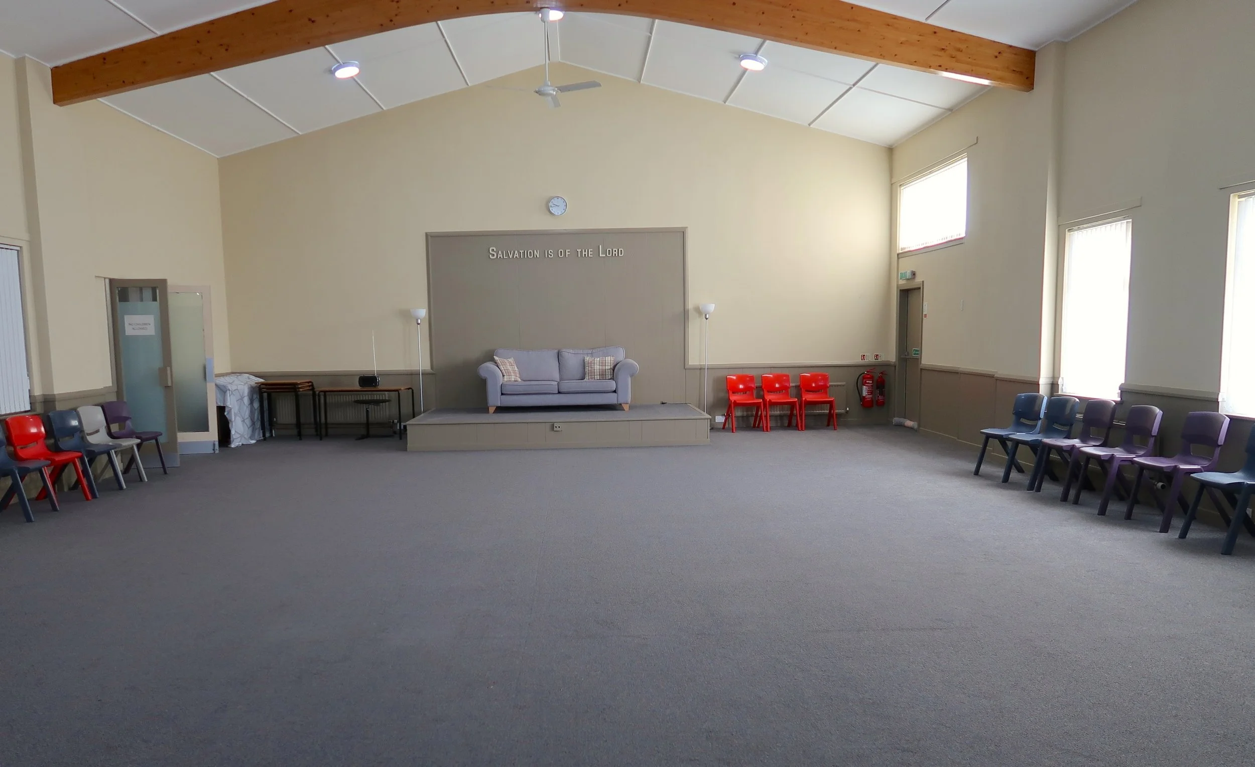 Empty multi-purpose room with chairs arranged along walls, a small stage with a gray sofa, and a sign that reads 'Salvation is of the Lord' on the wall behind the stage.