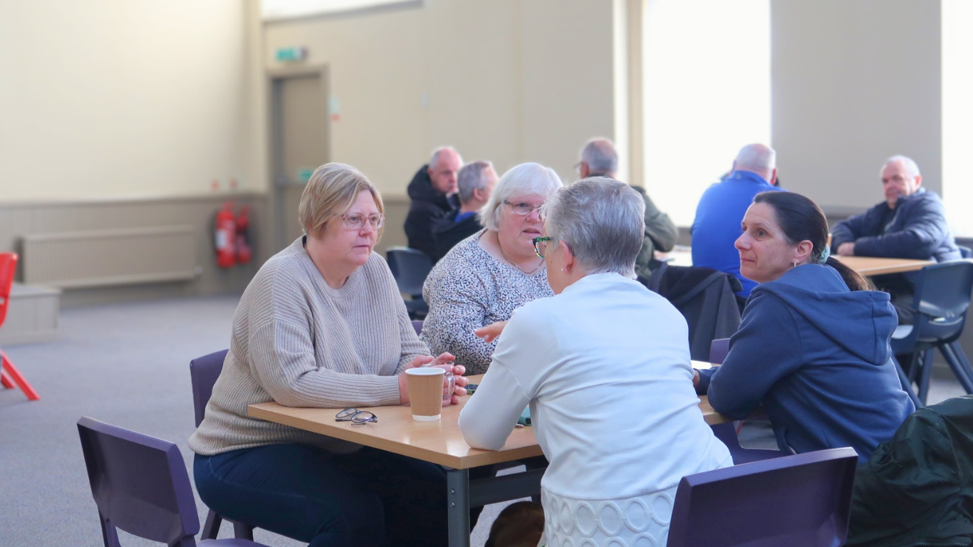 People sitting in groups at tables having conversations in a well-lit indoor space.