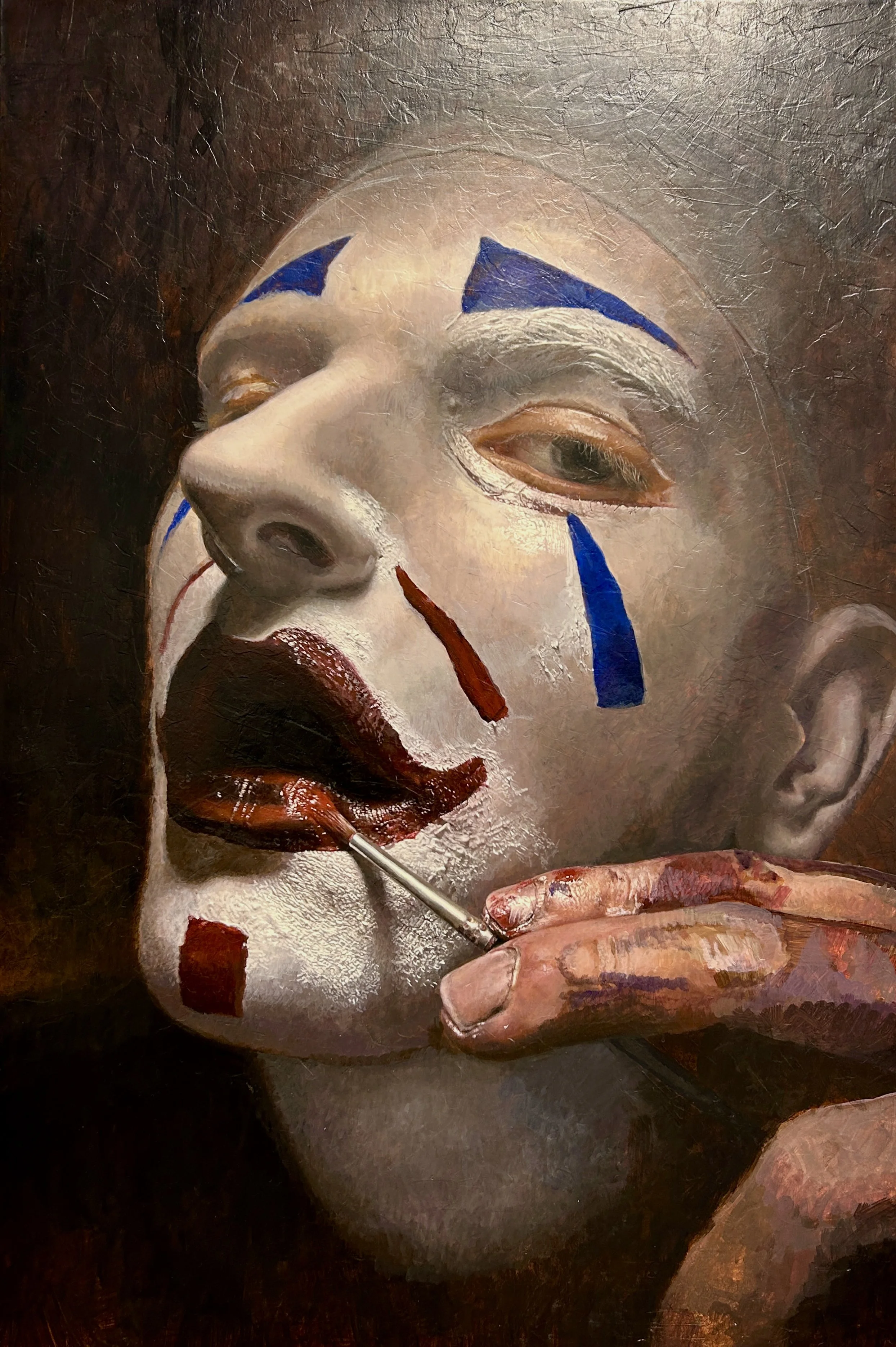 A person with clown makeup is applying red lipstick with a small brush. The makeup includes blue and white face paint, red on the lips, and white facial paint. The person's hand is visible holding the brush, and the background is dark.