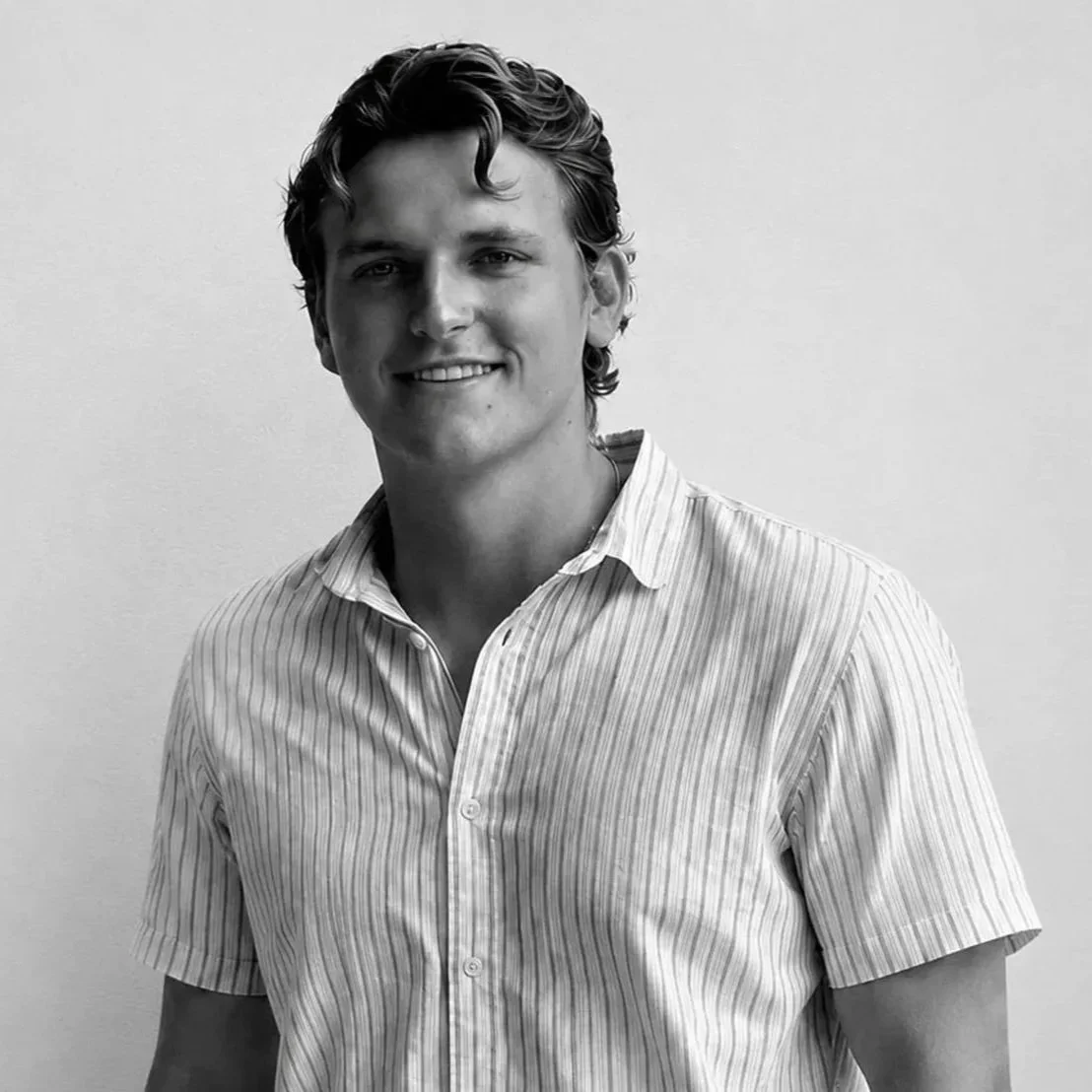 Black and white portrait of a young man with wavy hair, smiling, wearing a short-sleeved striped shirt.