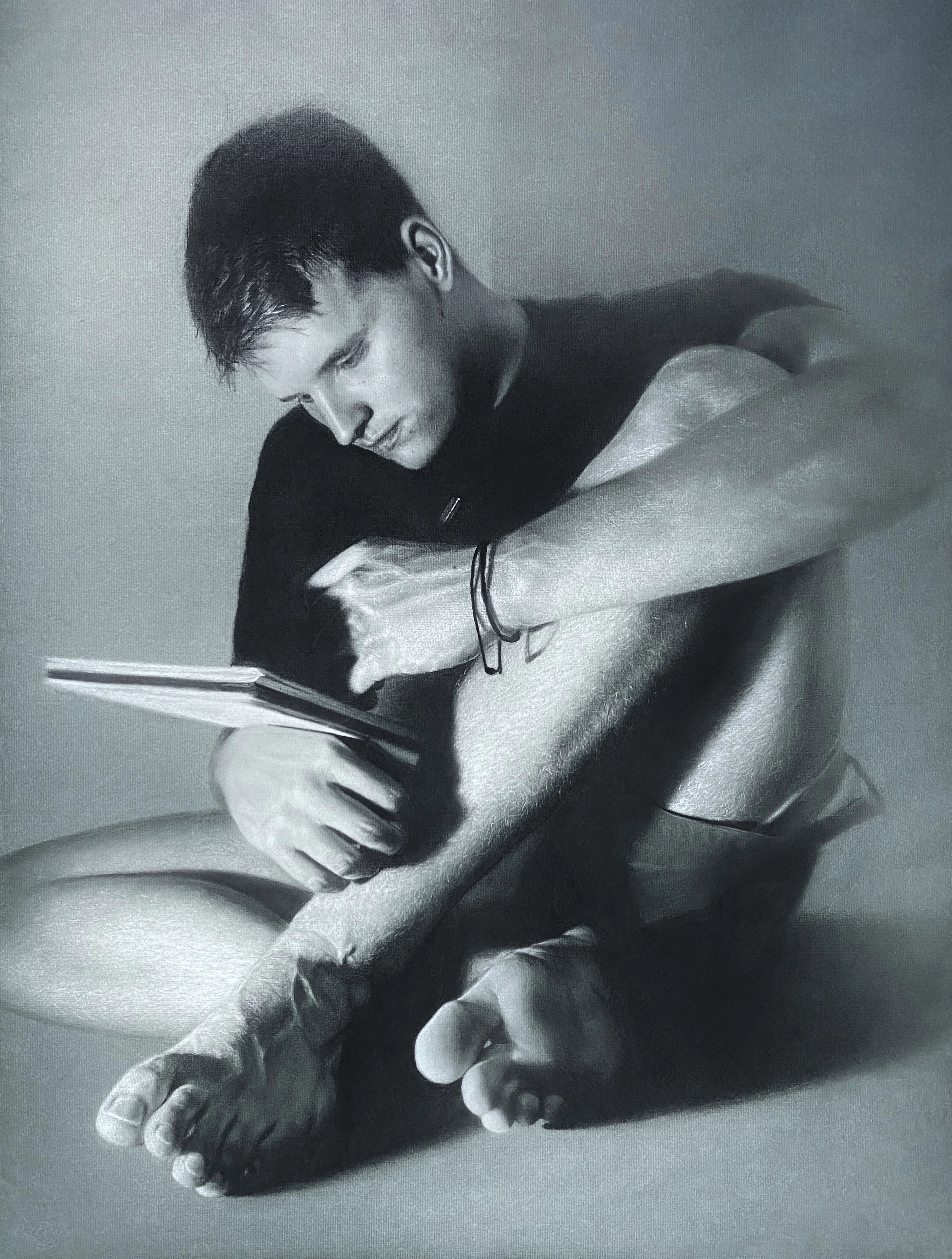 Black and white photograph of a young man with dark, wet hair hugging his knees, with closed eyes, sitting against a plain background.
