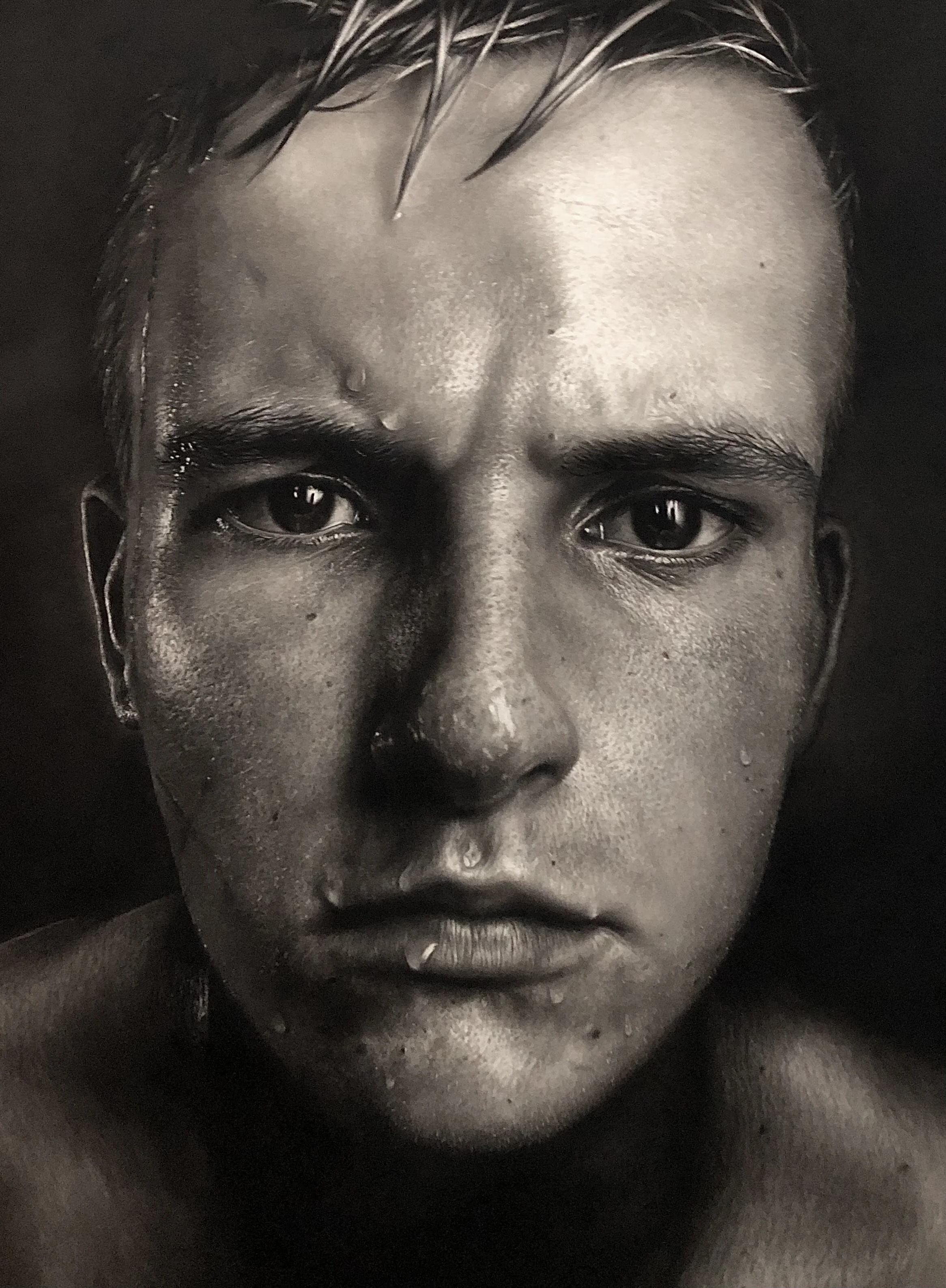 Close-up black and white photo of a young person's face with water droplets, dark eyes, and wet hair.