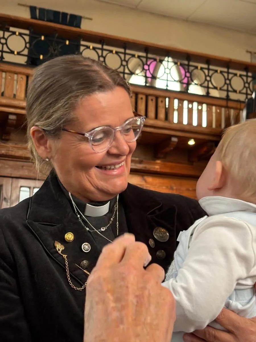A woman with gray hair, glasses, and a clergy collar smiling and holding a young child in a warm, wooden interior.