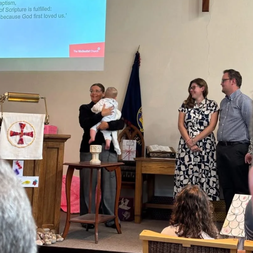 A woman holding a baby on stage during a church ceremony, with two people standing beside her. The stage features a wooden podium with a stained glass design and a flag in the background, with an open book on a desk. The church's wall is plain, and there are children and audience members visible in the foreground.