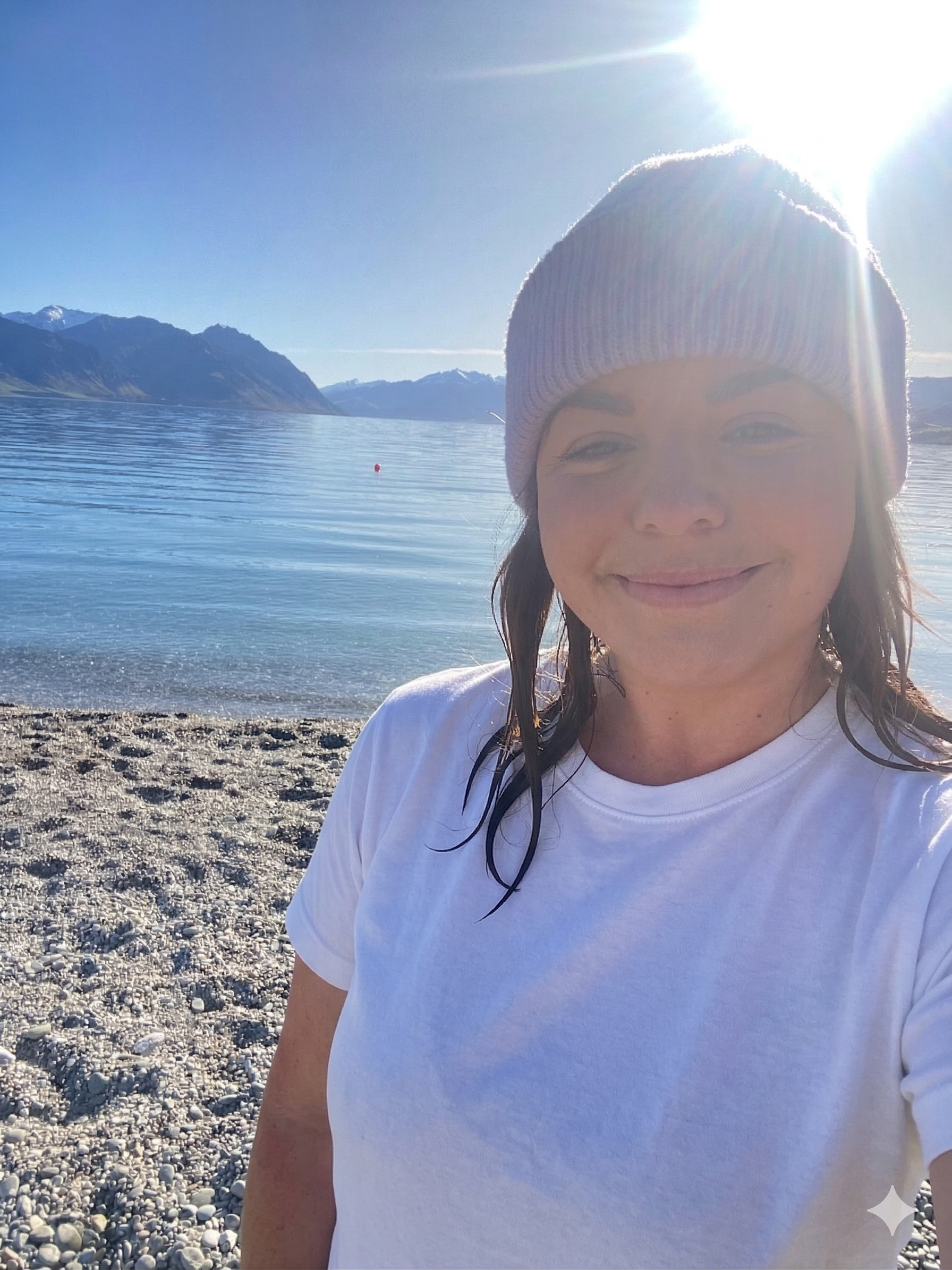 A woman taking a selfie on a pebble beach by a calm lake with mountains in the background, wearing a knit beanie and a white t-shirt, with bright sunlight overhead.