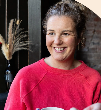 A smiling woman with curly hair wearing a red sweatshirt, holding a cup, standing indoors near a black door and a vase with dried plants.