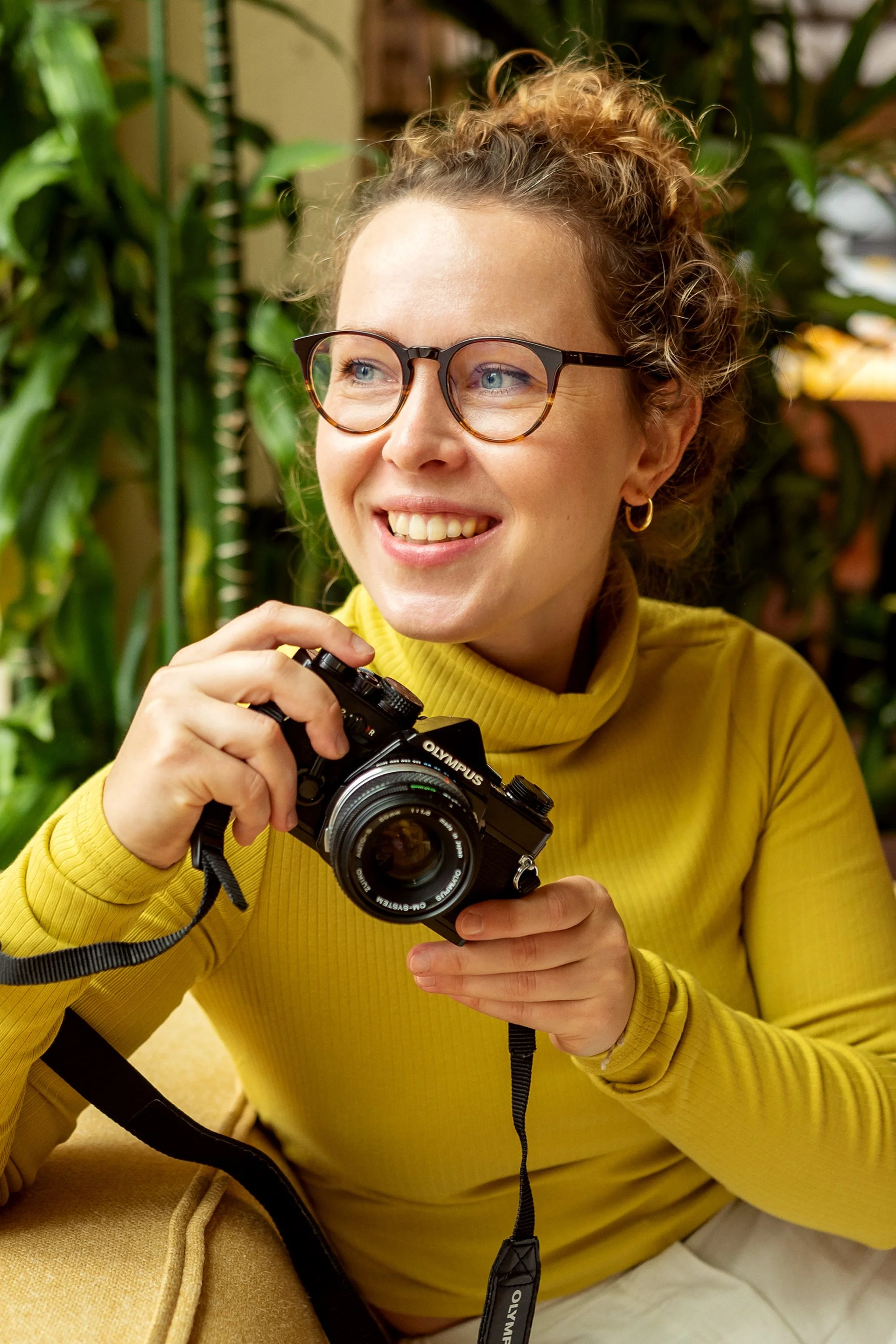 A woman with curly hair, glasses, and a yellow turtleneck sweater sitting indoors holding a camera and smiling.