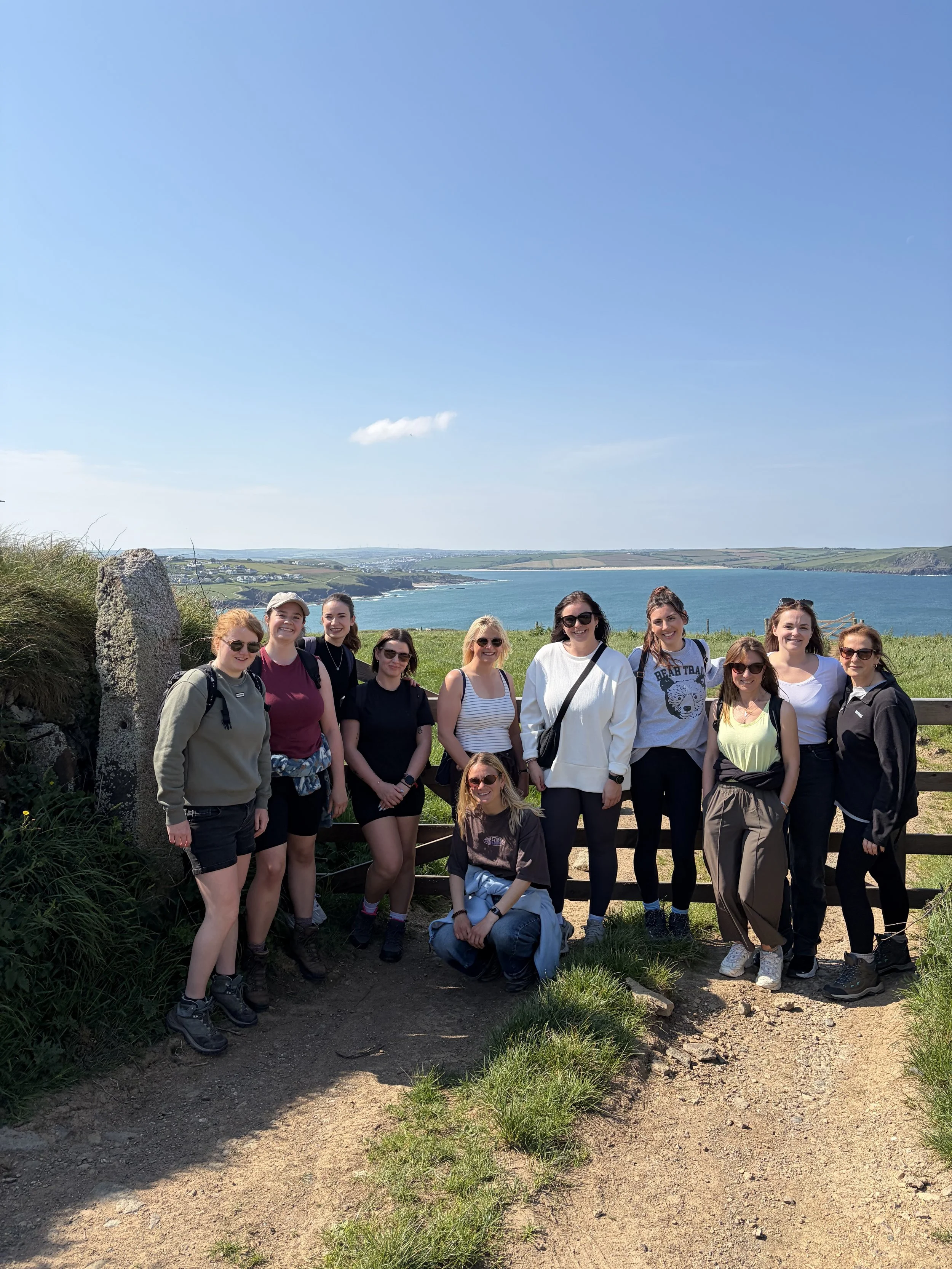 A group of women standing outdoors near a coastal area with a blue sky, green hills, and a body of water in the background.