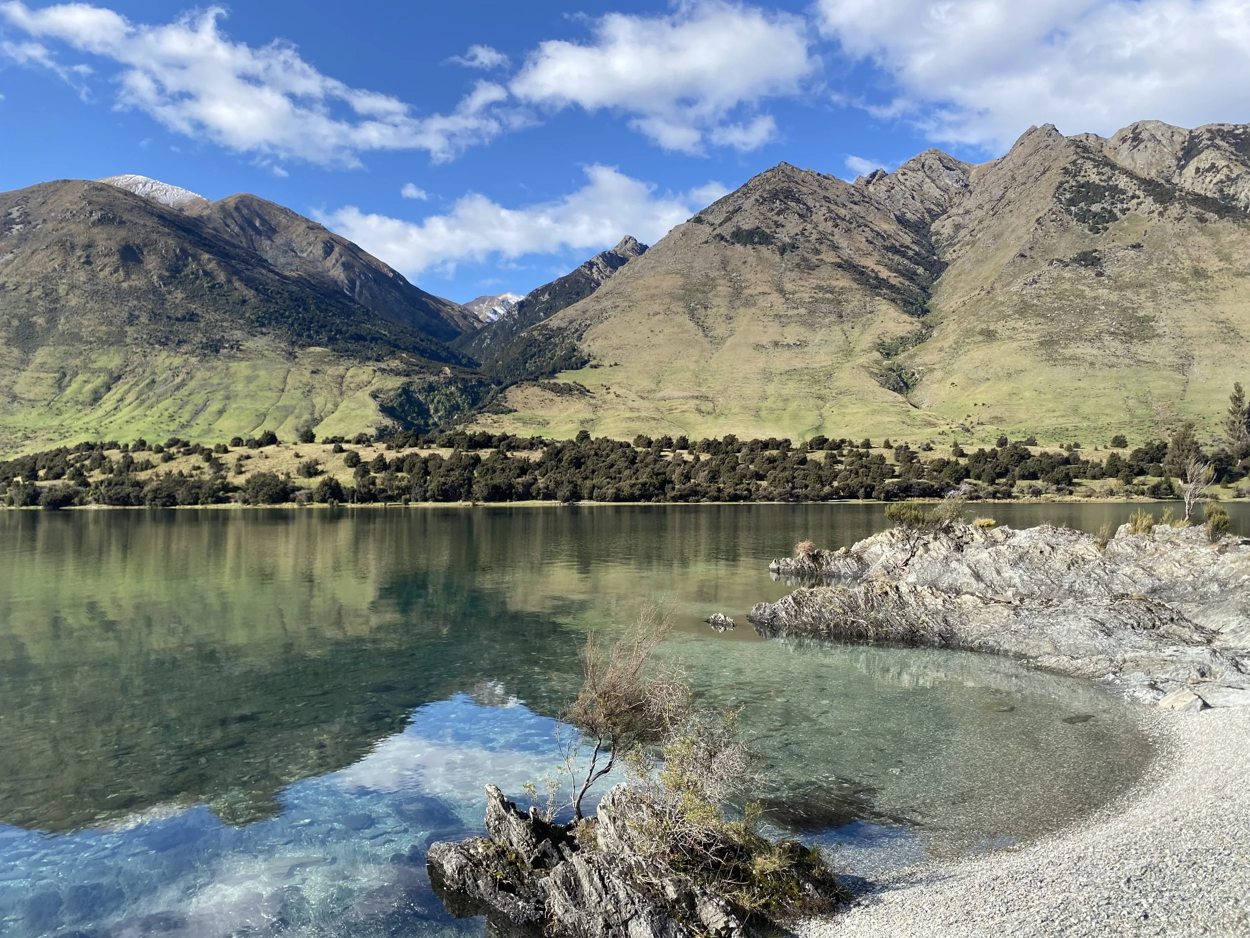 Scenic view of a calm lake with rocky shoreline, surrounded by green mountains and a blue sky with some clouds.