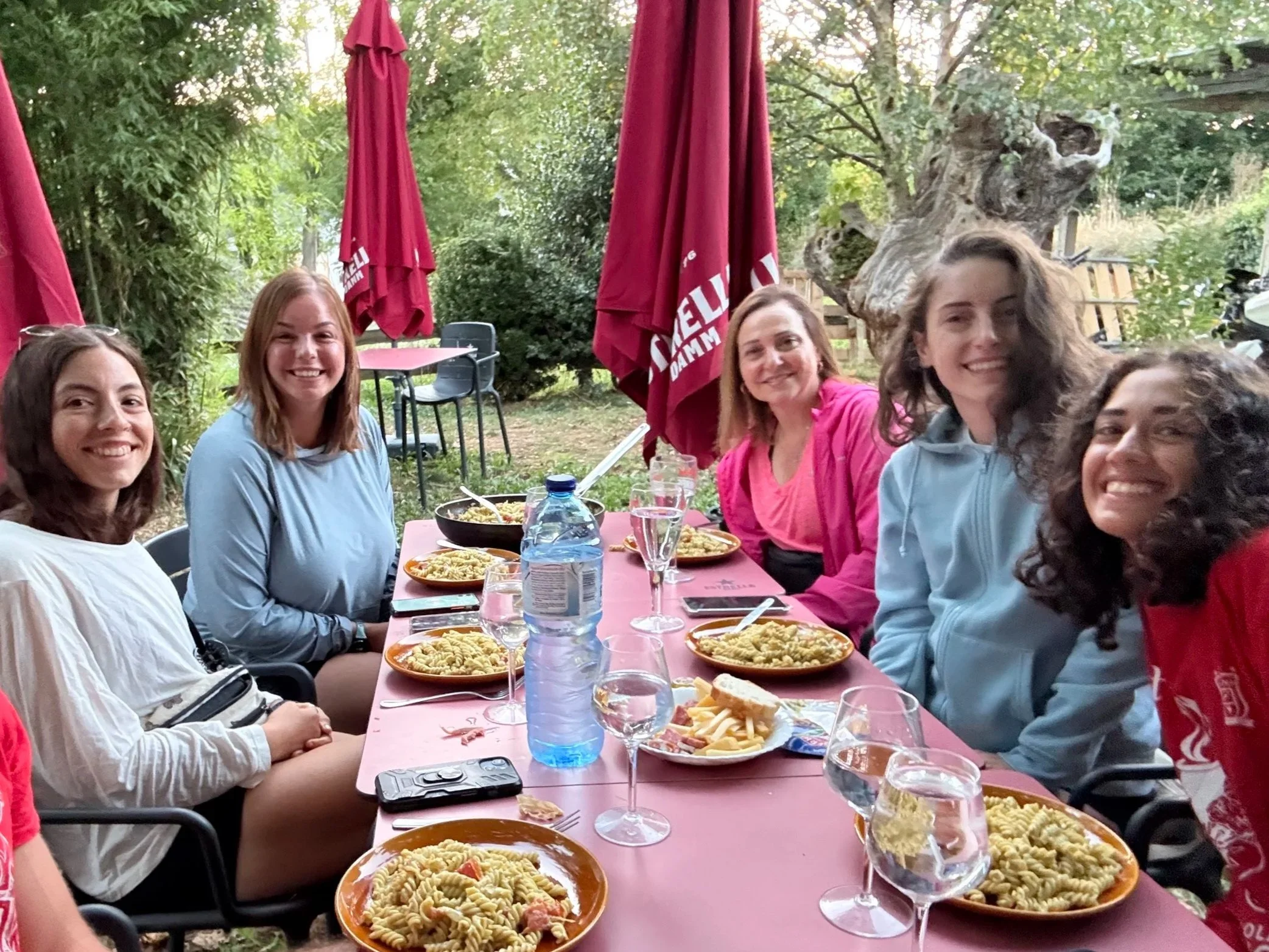 Six women sitting at a pink outdoor table with plates of pasta, empty glasses, water bottle, and cutlery, smiling, in a garden with trees and red umbrellas.