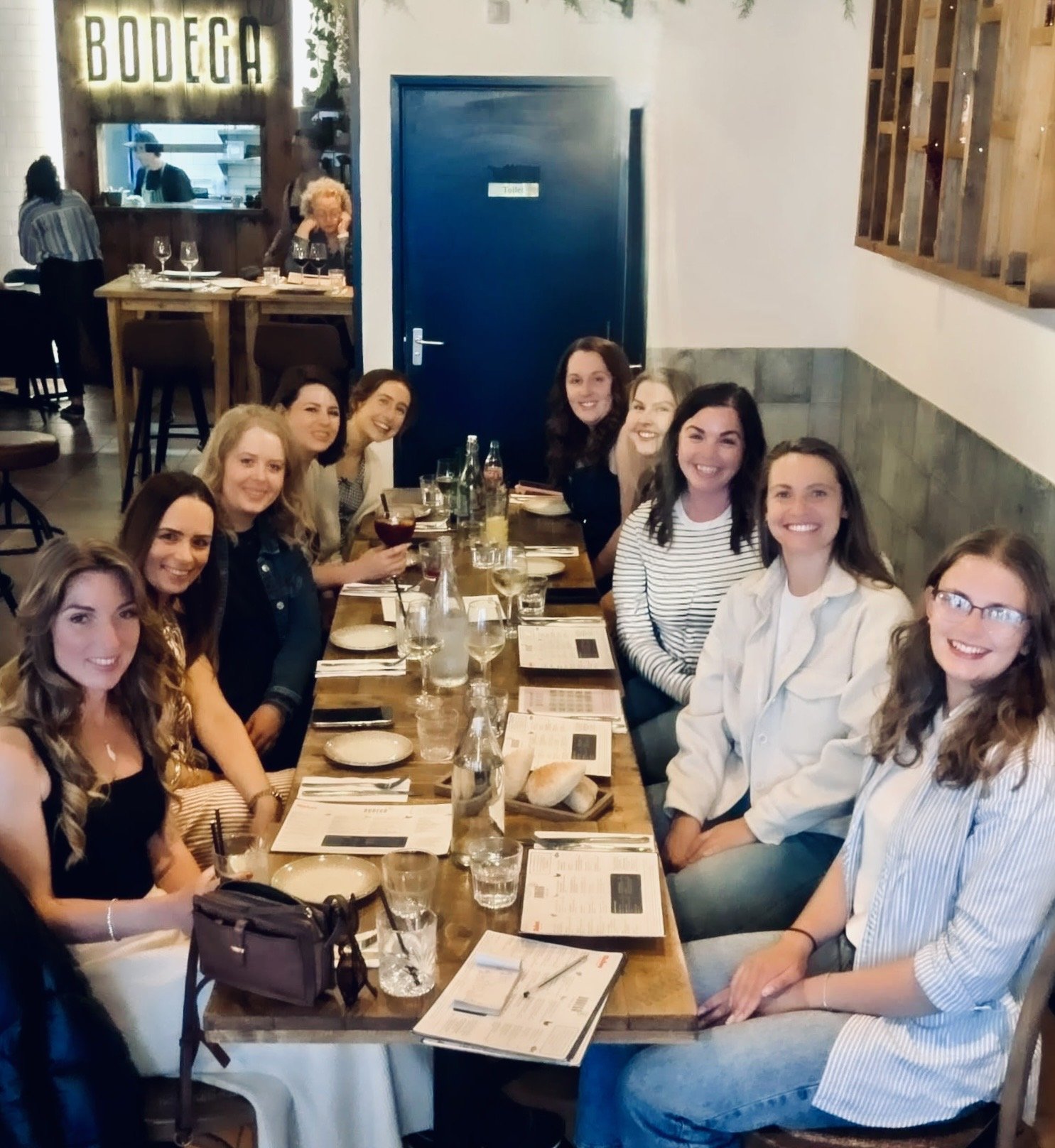 Group of women sitting around a long restaurant table, smiling, with menus, drinks, and bread. A blue door labeled "Toilets" is in the background. The restaurant has wooden and brick decor, with a sign reading 'BODEGA' on the wall.