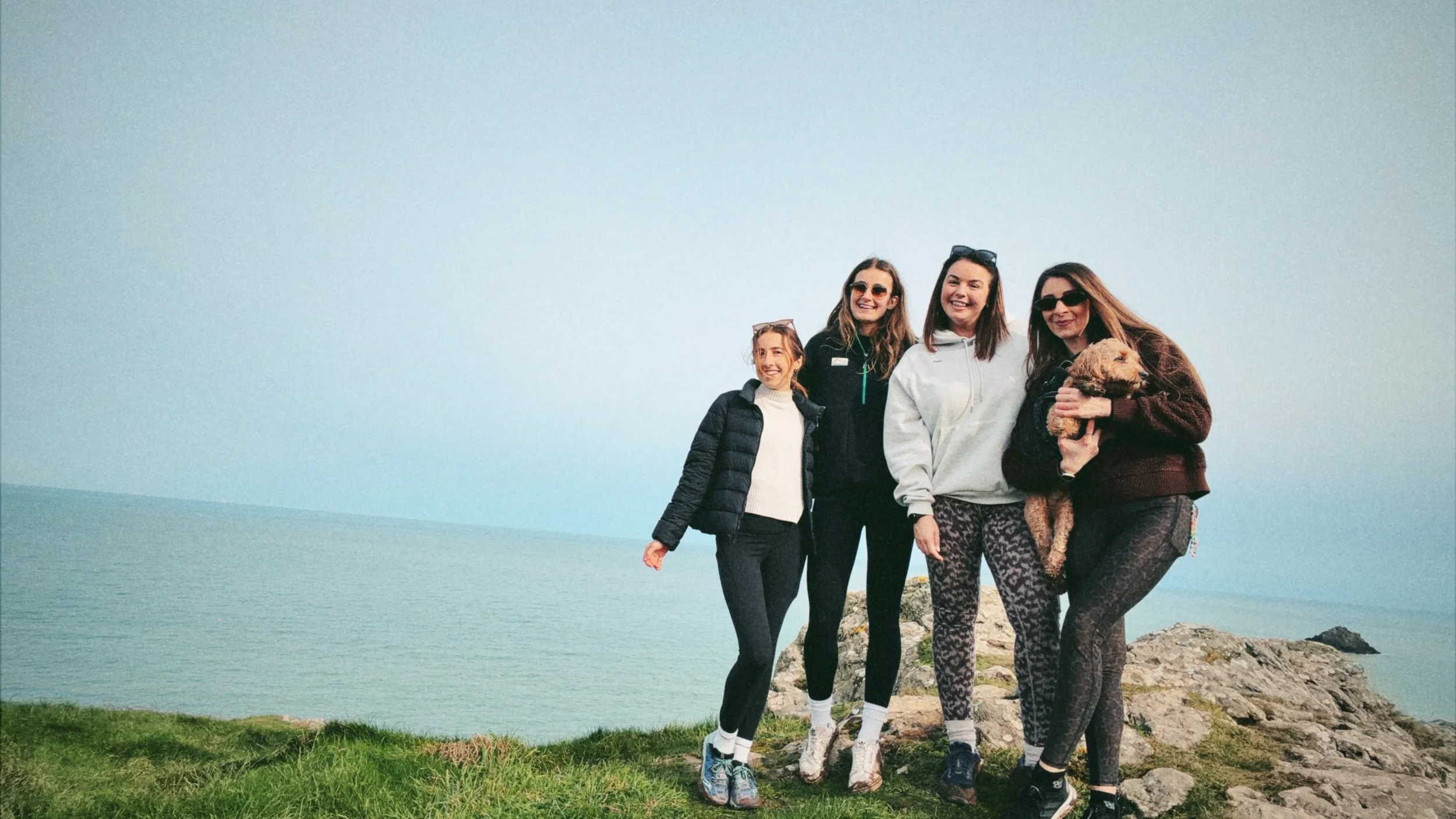 Group of four women and a dog standing on rocky terrain near the ocean, smiling and enjoying a walk.