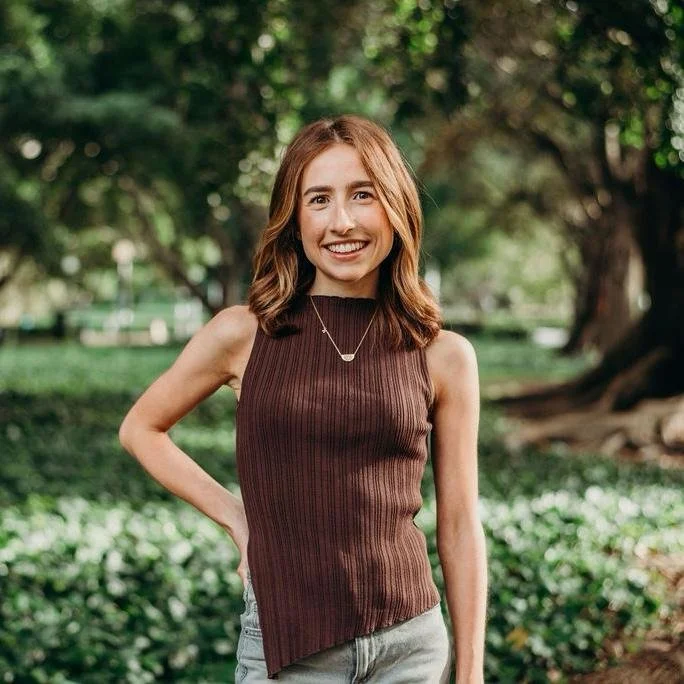 A young woman with shoulder-length brown hair, smiling, standing outdoors in a park with greenery and trees in the background.