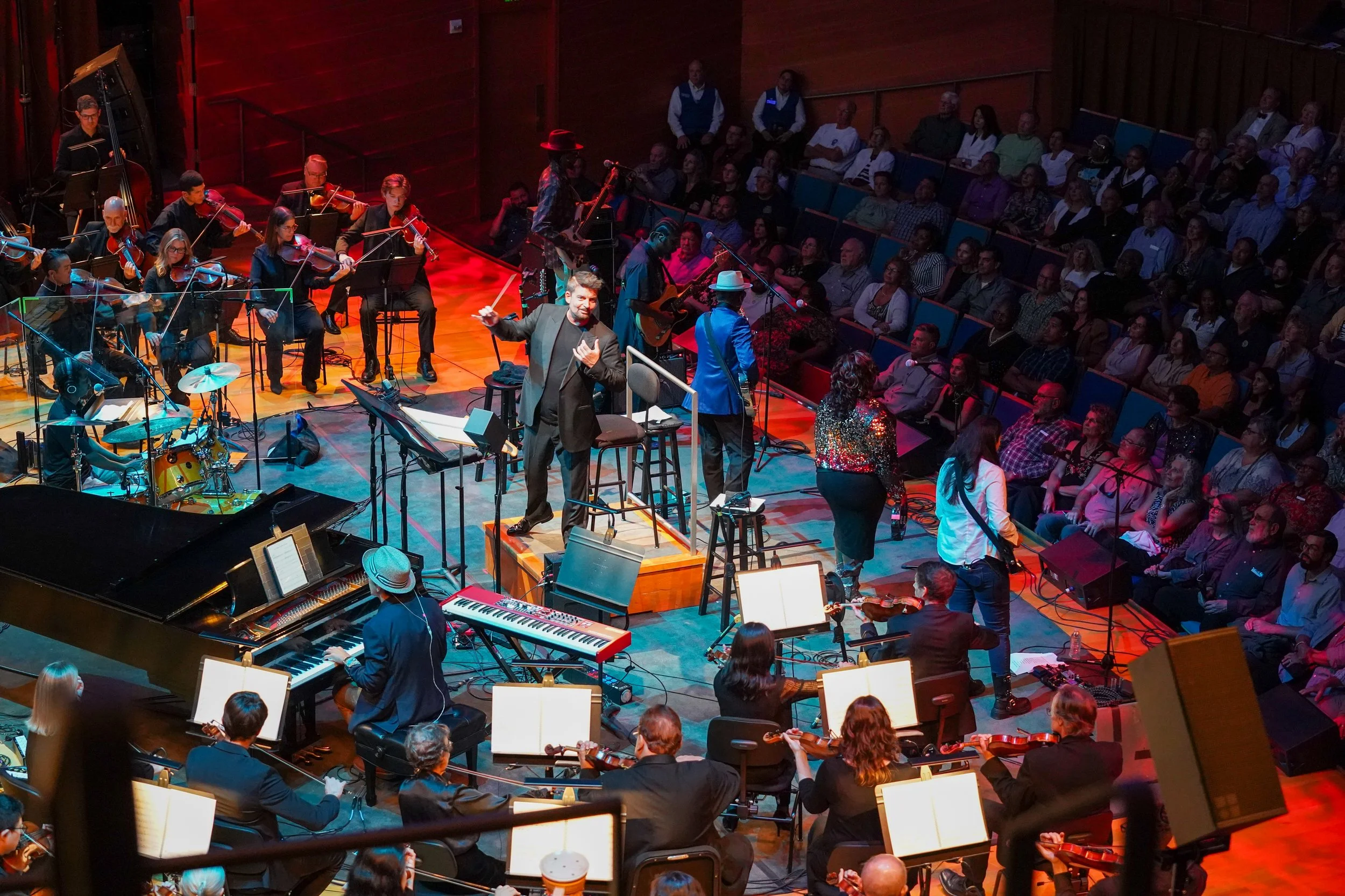 Orchestra performing on stage with conductor, in front of audience in concert hall.